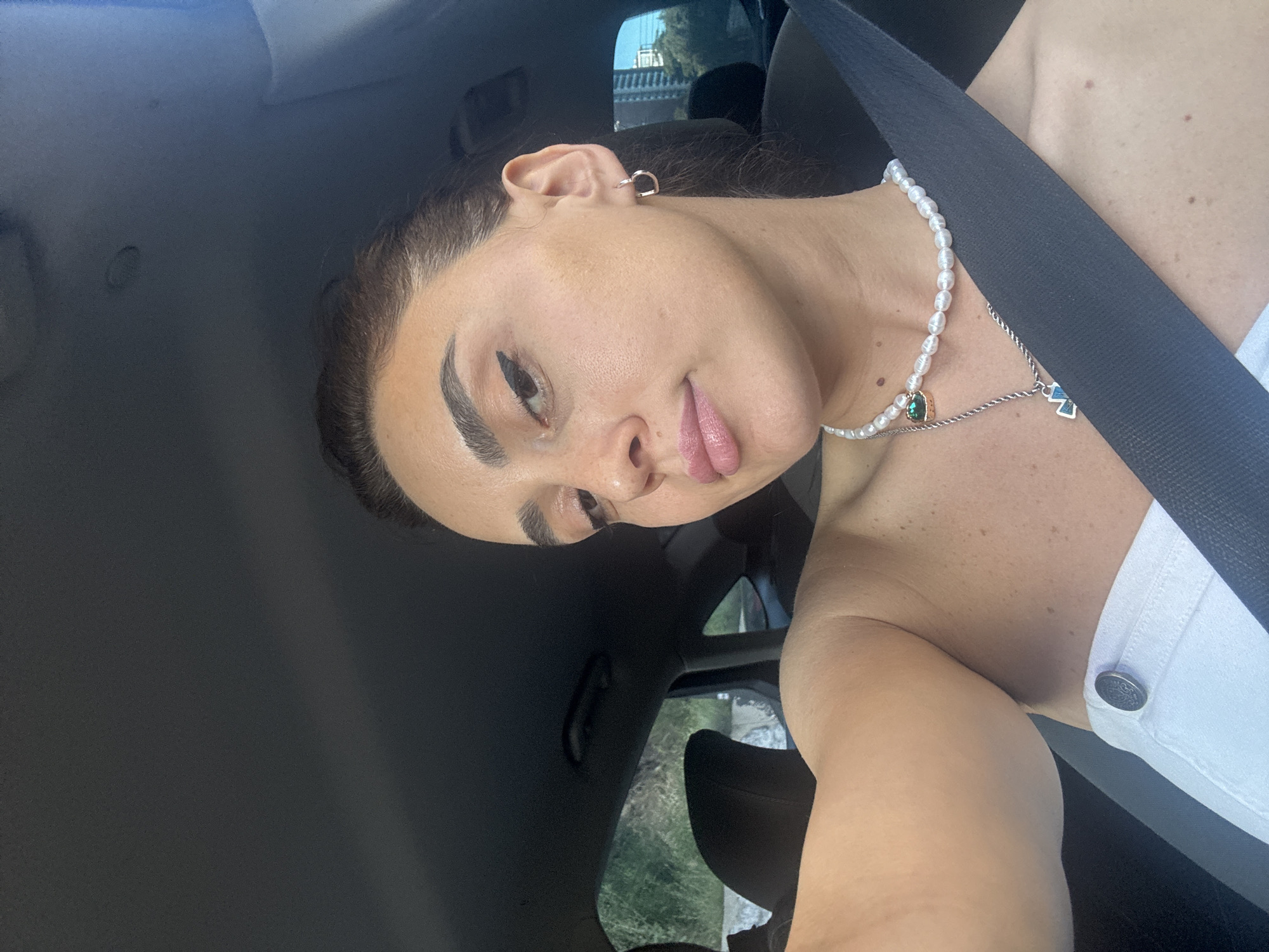 A woman is taking a selfie in the passenger seat of a car, with her arm extended towards the camera. She has brown hair pulled back and is wearing a white top, a pearl necklace with a green pendant, and another silver necklace with a blue pendant. She also has a gold hoop earring in her right ear. The interior of the car is visible, including a black seat and a seatbelt. Outside the car, trees and buildings can be seen, suggesting an urban or suburban environment. The image does not provide enough information to determine the specific location within Tbilisi, Georgia.