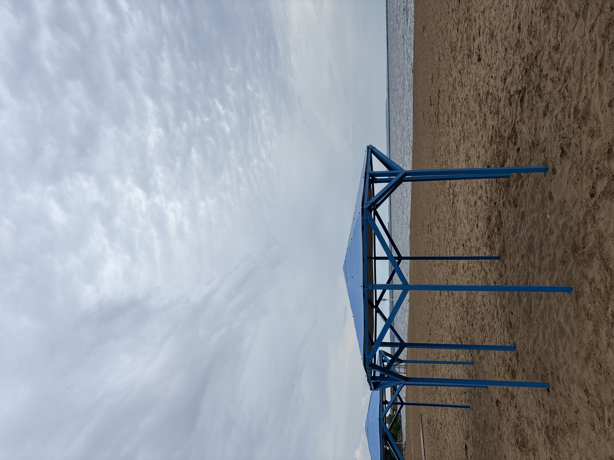 The image depicts a tranquil beach scene, likely near the Kama River or Votkinsk Reservoir in Chaikovsky, Russia. The primary subjects are several blue, metal shade structures or cabanas, positioned along a sandy shore beside a body of water.
The scene is dominated by a wide expanse of light brown sand, textured with small undulations and footprints, stretching from the foreground towards the water's edge. To the right, a calm, grey-blue body of water meets the horizon. Above, the sky is overcast with a blanket of soft, diffuse grey and white clouds, indicating a cloudy or partly cloudy daytime, possibly under subdued sunlight.
The blue structures are made of metal, featuring a minimalist, angular design with vertical posts and horizontal beams supporting a flat, possibly tarp-covered, roof. Several of these structures are visible, receding into the distance along the shoreline, suggesting a series of beach amenities. No people are visible, implying the beach is empty or sparsely populated, perhaps during an off-season or a quiet part of the day. There is no discernible activity taking place. No visible text is present in the image.
