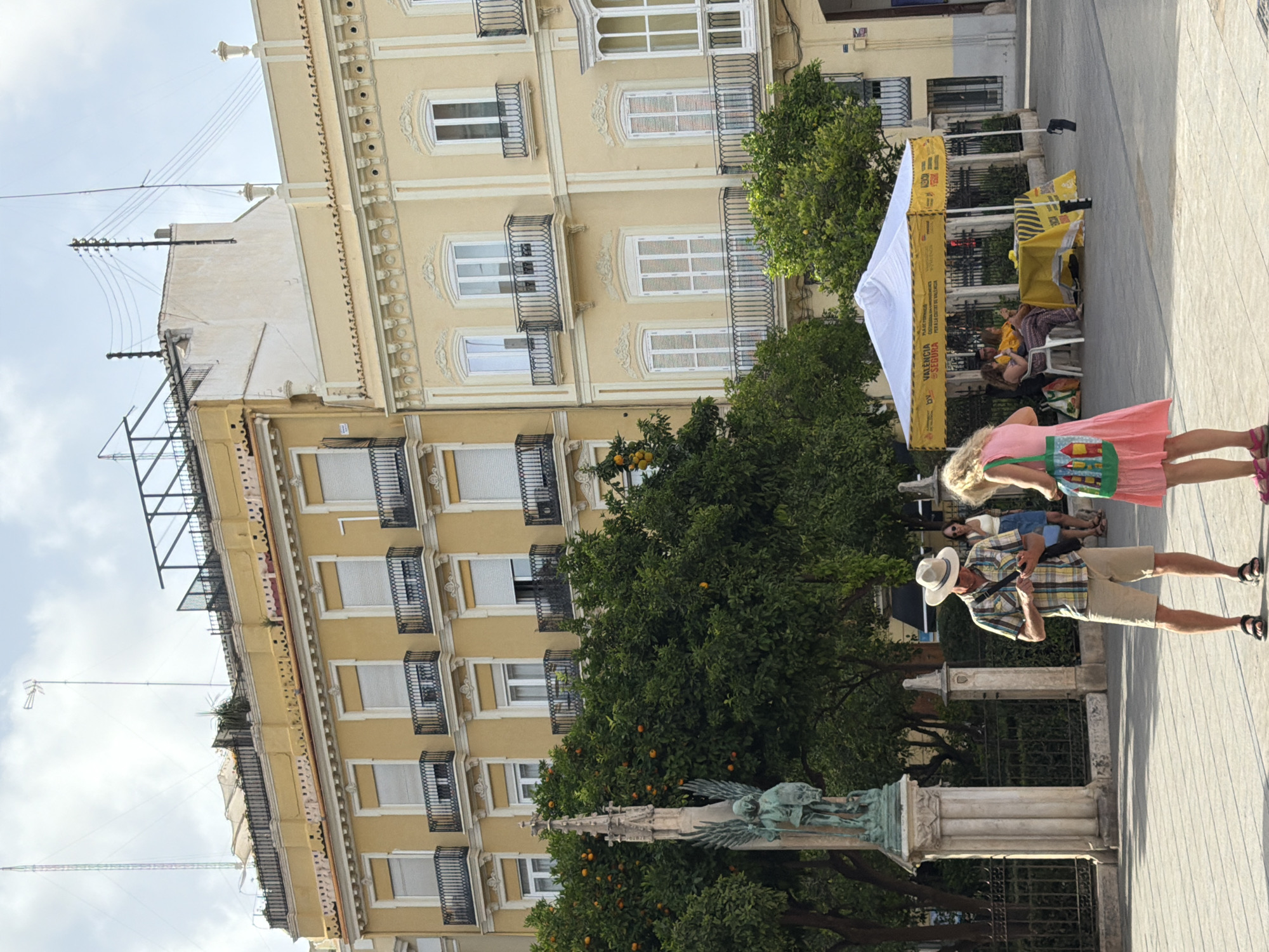 In an outdoor plaza, two people stand near a monument and trees laden with fruit. A man in a plaid shirt and shorts looks at his phone, while a woman in a pink dress stands nearby. In the background, a white tent with yellow accents provides shade, with several people seated under it. Yellow buildings with ornate balconies line the plaza, and a prominent antenna rises into the sky above them. The scene is bathed in natural light under a cloudy sky.