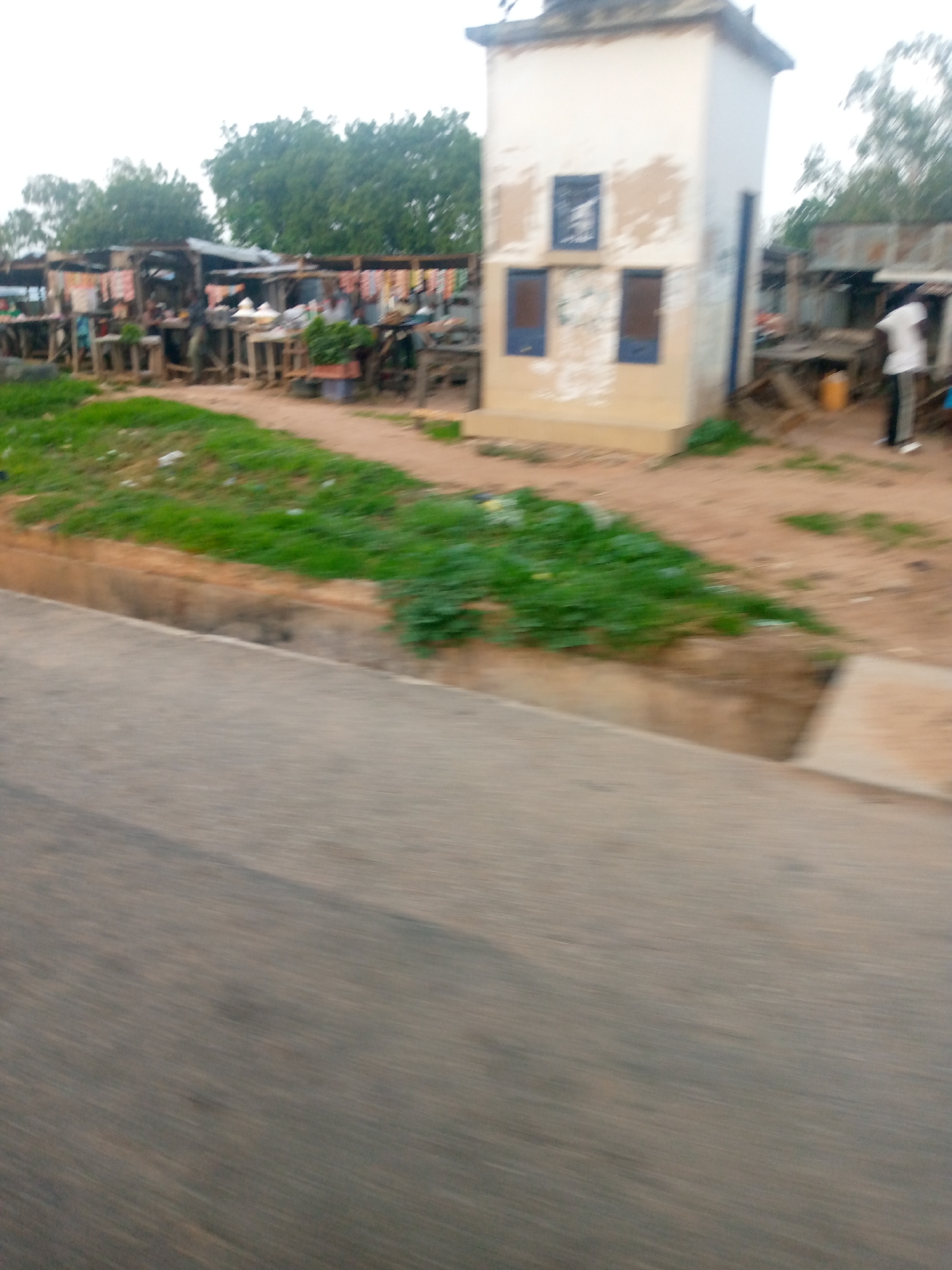 The image captures a street-level view of a bustling marketplace in Jalingo, Nigeria, likely situated along a main thoroughfare, evidenced by the asphalt road in the foreground and the presence of a drainage channel. A small, cuboid building with white and beige paint, possibly a utility booth or small office, stands prominently to the right, featuring blue-framed window openings. Adjacent to this structure and extending to the left is a row of open-air market stalls constructed from wood and corrugated metal roofing. These stalls display various goods, including textiles hanging from racks and produce or other items arranged on tables. Several individuals are visible within the market area, some engaged in commercial activities, and one person in a white shirt and dark pants stands near the booth. The environment is characterized by dirt ground, patches of green grass, and scattered debris. The overall impression is of a vibrant, everyday scene in a Nigerian urban setting.