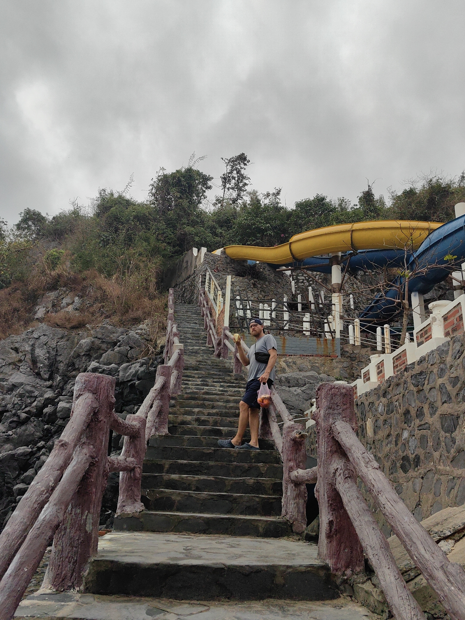 The image depicts a man standing on a long set of concrete stairs with wooden railings, located next to a rocky cliff and a water slide structure. The man is wearing a gray t-shirt, dark shorts, and a baseball cap. He is holding a banana in his right hand and a plastic bag containing fruit in his left. Behind the man is a water slide structure with a yellow and blue slide. The stairs are flanked by a stone wall on the right side. The environment includes lush vegetation on the top of the cliff and a gray, overcast sky. The location is in the city of Hải Phòng, Vietnam.