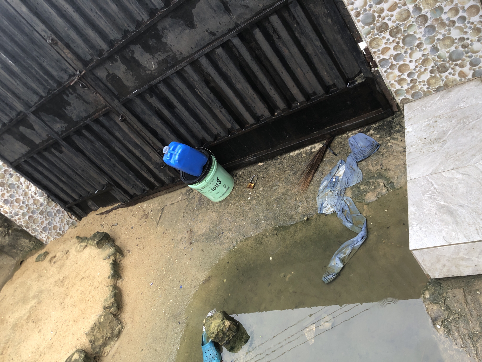 The image depicts an outdoor scene in Ado, Nigeria. In the foreground, a puddle of murky water covers a portion of a concrete surface. Discarded items, including a blue plastic bottle and a piece of blue fabric, are submerged in the water or lying nearby. To the left of the puddle, a light blue sandal rests on a rock. Behind the puddle, a green bucket with a black lid is visible, topped with a blue plastic container. A broom lies on the ground next to the bucket. In the background, a dark metal gate with horizontal slats dominates the right side of the frame, showing signs of rust and wear. The ground adjacent to the gate is covered with light brown sand and decorated with circular patterned tiles in earthy tones. The overall impression is one of an unkempt outdoor area with evidence of recent rain or standing water.
