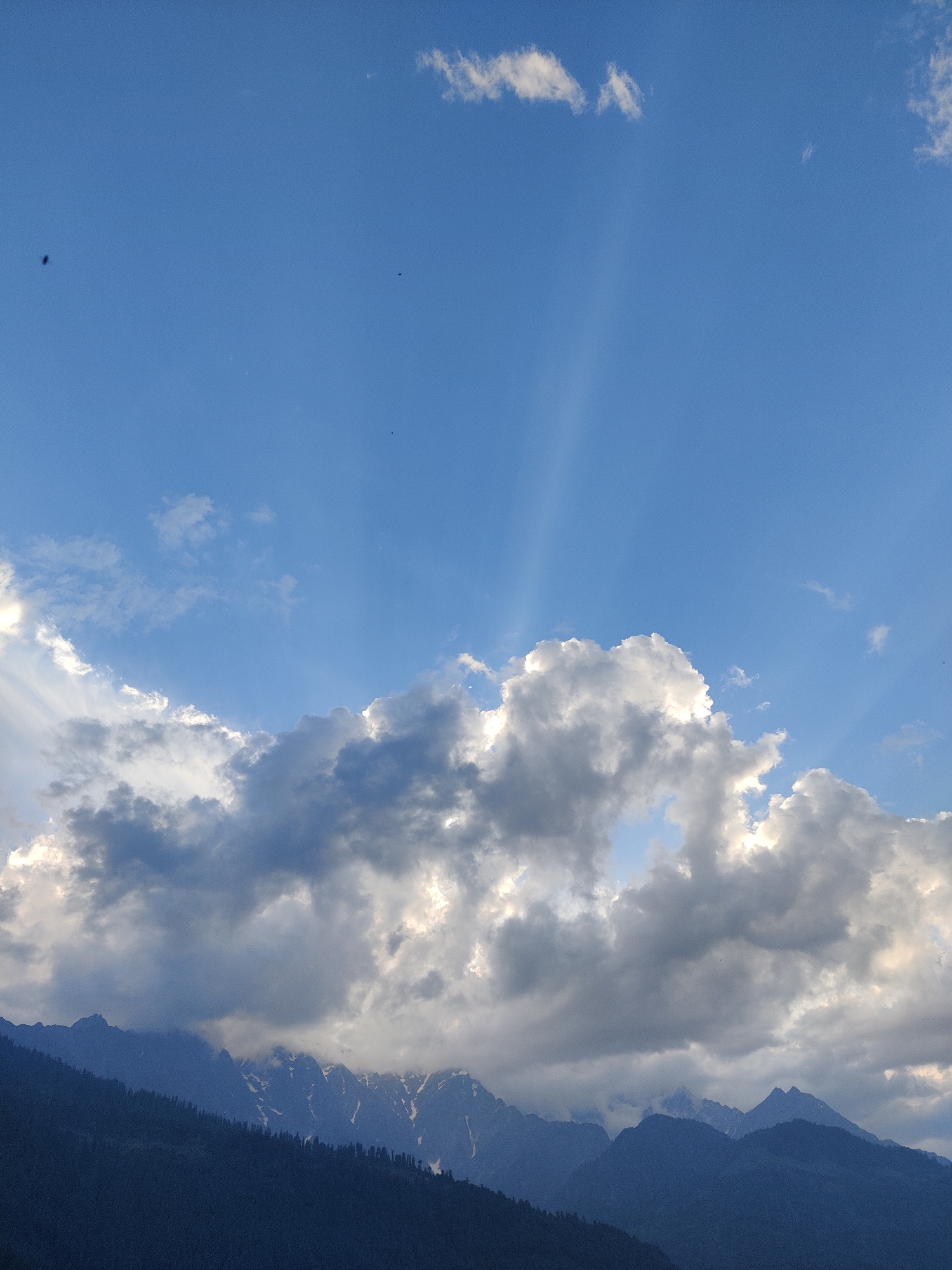 The image captures a majestic mountain landscape at dusk, likely in the Vashisht region of India, given the style of the mountains and the presence of snow-capped peaks. The foreground is dominated by the dark, silhouetted slopes of heavily forested mountains. Above them, the rugged peaks of even higher mountains pierce through the lower cloud cover, revealing patches of snow on their summits. A dramatic sky fills the upper half of the image. Large, billowing clouds, some dark and heavy with the promise of rain and others illuminated by the fading sunlight, create a dynamic visual effect. Rays of sunlight, known as crepuscular rays, are visible, fanning out from behind the clouds, suggesting the sun is low on the horizon. The overall atmosphere is one of natural beauty and awe, typical of mountainous regions at twilight. There are no visible people or man-made structures in the image, focusing solely on the natural environment.