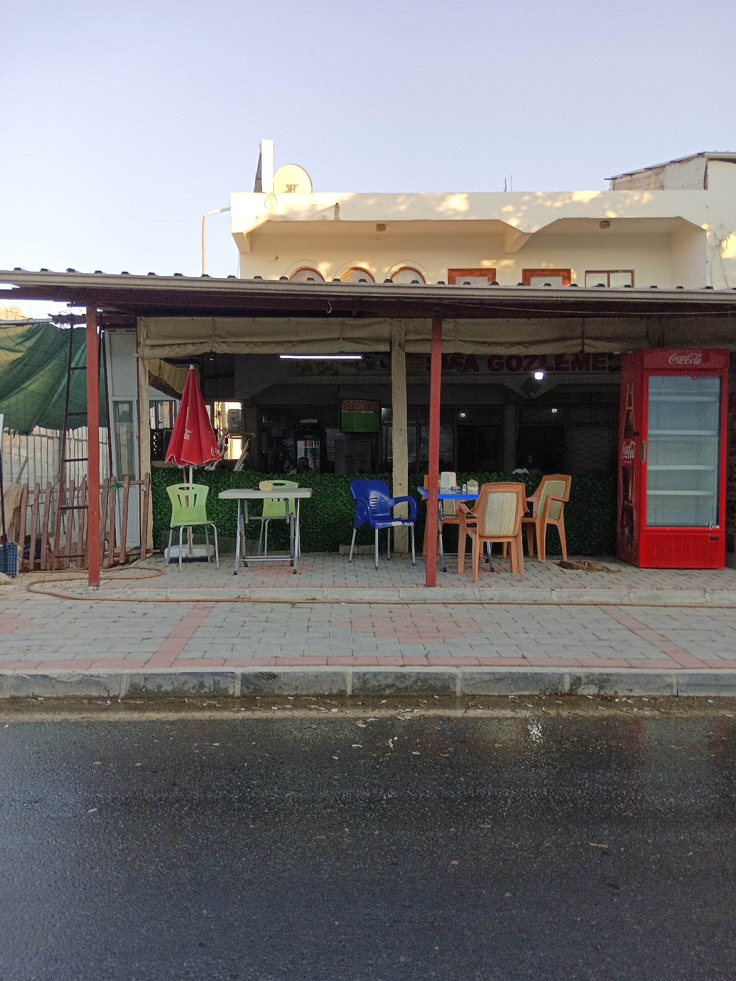The image depicts the exterior of a small establishment, likely a cafe or restaurant, on a street in Gazipaşa, Turkey. In the foreground, a wet asphalt road runs parallel to a paved sidewalk. The sidewalk is constructed with interlocking gray pavers bordered by red-toned rectangular pavers. Beyond the sidewalk, a covered outdoor seating area is visible, supported by red metal posts. Several tables and chairs are arranged in this area. To the left, a red-striped umbrella stands near a small table with two light green chairs. Further to the right, a blue chair sits beside a table with four tan chairs. A prominent red Coca-Cola branded beverage cooler is positioned at the far right of the seating area. The establishment's facade features a light-colored awning and signage that appears to read "FA GÖZLEM...". Above the awning, the upper floors of a building are visible, with a satellite dish on the roof. The environment appears to be outdoors, with clear skies and some dappled sunlight suggesting late afternoon or early morning. No people are clearly visible, but the presence of tables and chairs indicates it is a public space.