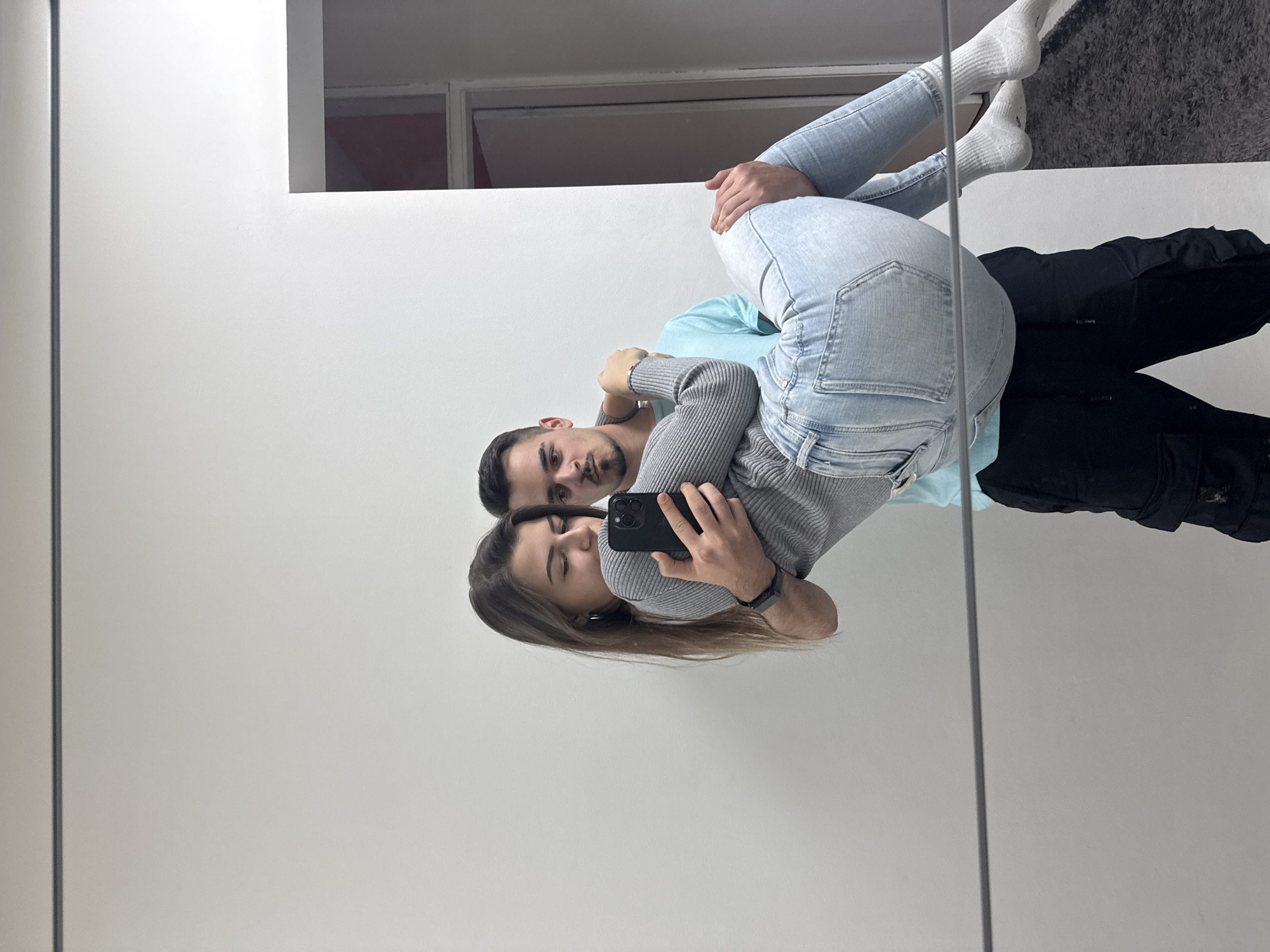 A young adult couple is captured in what appears to be a mirror selfie, with the image rotated ninety degrees clockwise. The woman, with long brown hair, is in the foreground holding a black smartphone with her right hand, aiming it towards the mirror's reflection. She wears a grey ribbed long-sleeve top and light blue jeans, paired with white socks. Her expression is a slight smile or smirk, with her eyes focused on the phone screen.
Behind her, the man has his arms wrapped around her, seemingly in an embrace. He has short dark hair and a light beard, with a neutral expression looking directly towards the phone's camera. He is wearing a light blue t-shirt. The reflection reveals his lower body clad in black pants. A black wristwatch is visible on his left wrist.
The setting is an indoor space, brightly illuminated by unseen light sources. The couple is reflected in a large, clear mirror. In the upper right corner of the reflection, a darker, rectangular area suggests a window or opening to another room, possibly with a darker interior or indicating a nighttime view. The prominent background behind the couple is a clean, white wall or surface. A thin, dark vertical line runs along the left side of the frame, possibly an edge or frame of the mirror or a structural element of the room. The scene takes place in Sofia, Bulgaria. There is no visible text in the image.