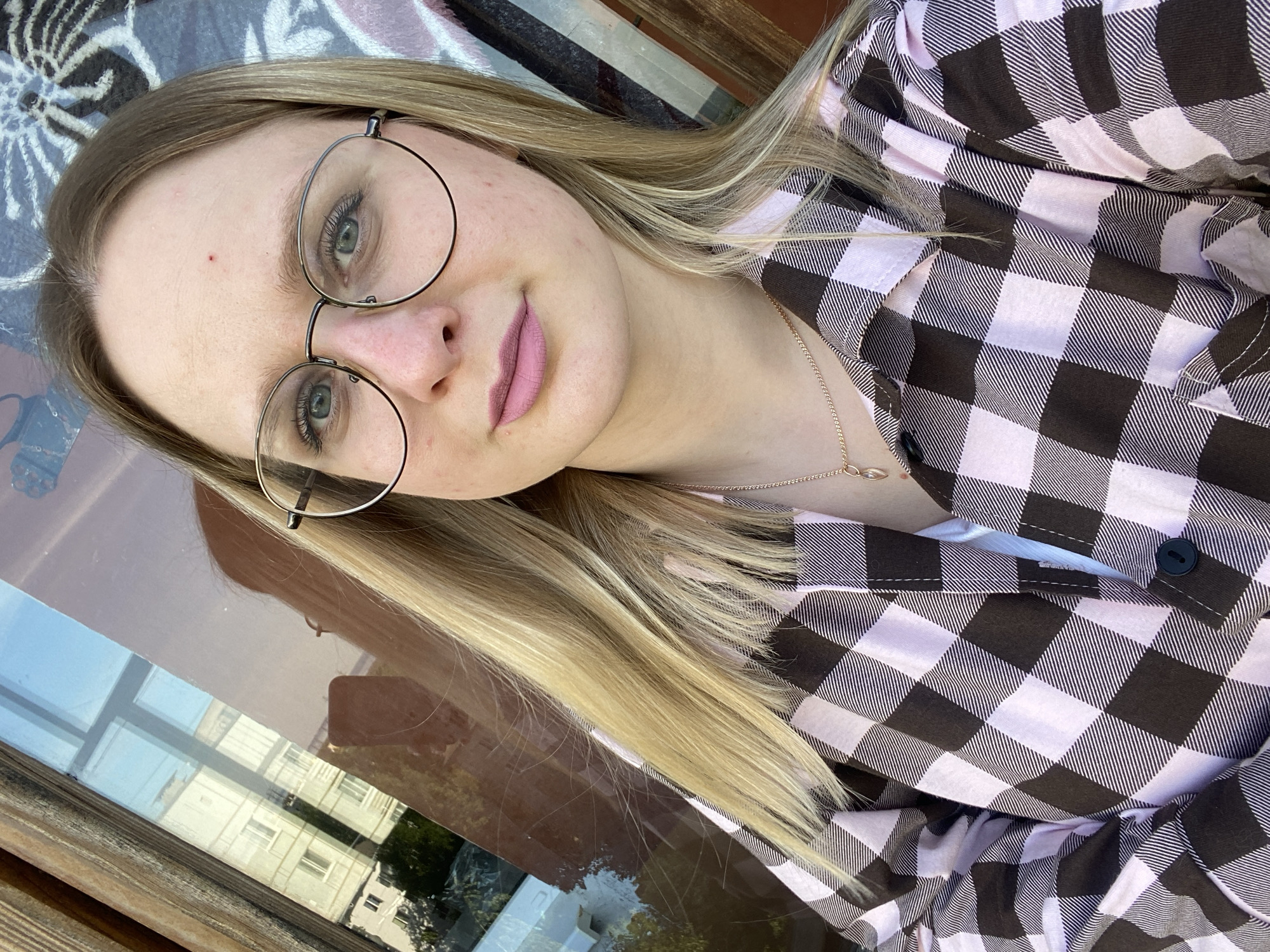 A medium shot depicts a fair-skinned young woman with blond hair wearing round, metal-rimmed glasses and a pink and black checkered shirt. She is adorned with a gold necklace. The background reveals a cityscape reflected in a window, implying an urban setting, potentially in Unknown, Unknown. The woman appears to be in an outdoor location near the window, suggesting a street-level view.