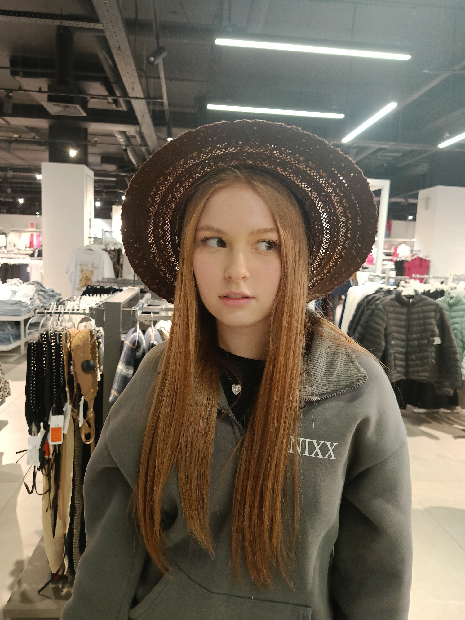 A young woman with long auburn hair is wearing a brown woven hat and a gray zip-up sweatshirt with the word "NIXX" printed on the left side of her chest. She is looking off to her left, and her expression is neutral. She is standing in a clothing store with racks of clothing visible in the background. The lighting is bright and fluorescent. The location is Izhevsk, Russia.