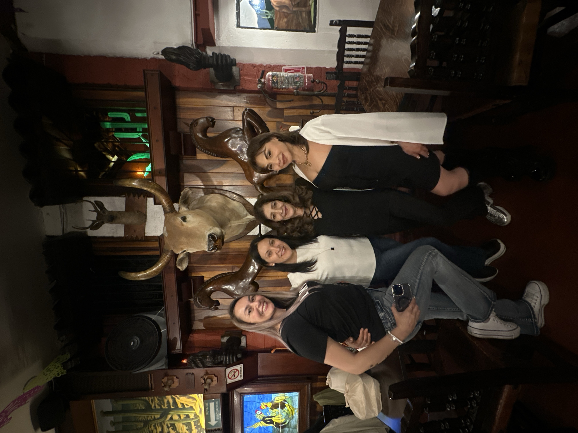 Four women are posing for a photo in what appears to be a restaurant or bar. The women are dressed in casual attire: tops, jeans, and sneakers. The room features dark wood paneling, mounted animal heads, and decorative objects. The location may be in Mexico City, Mexico.