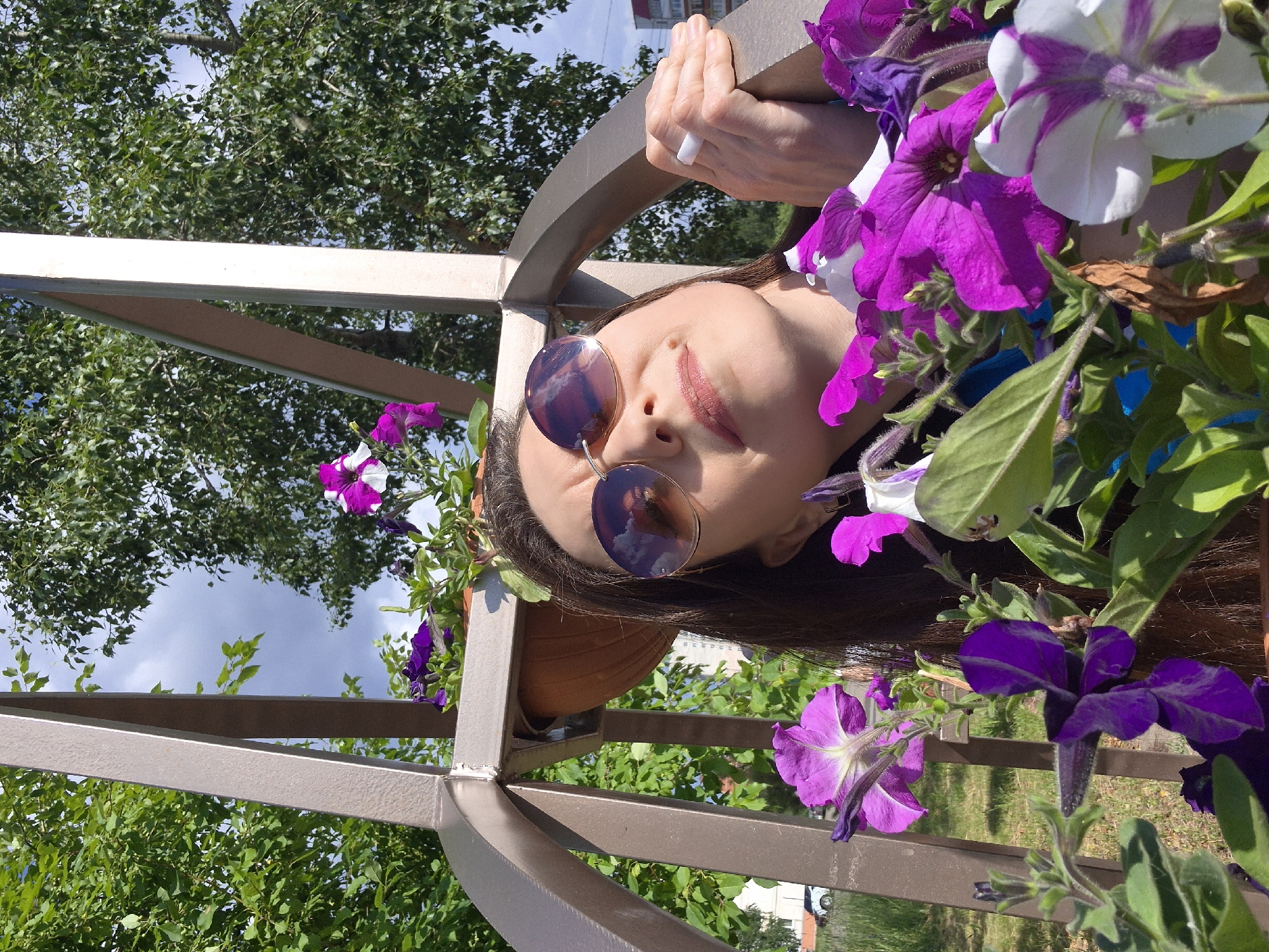 A medium shot captures a woman with dark hair and sunglasses posing outdoors amidst a profusion of purple and white petunias. She is positioned behind a metal structure, possibly part of a decorative garden feature or play equipment. Her head is tilted upwards, and she is looking towards the sky, her right hand resting on the metal structure. The background reveals lush green trees and a cloudy sky, suggesting a summer day. A distant building and a parked car are visible on the left side of the frame. The image appears to be taken in an urban park or garden area in Chaikovsky, Russia, given the context.