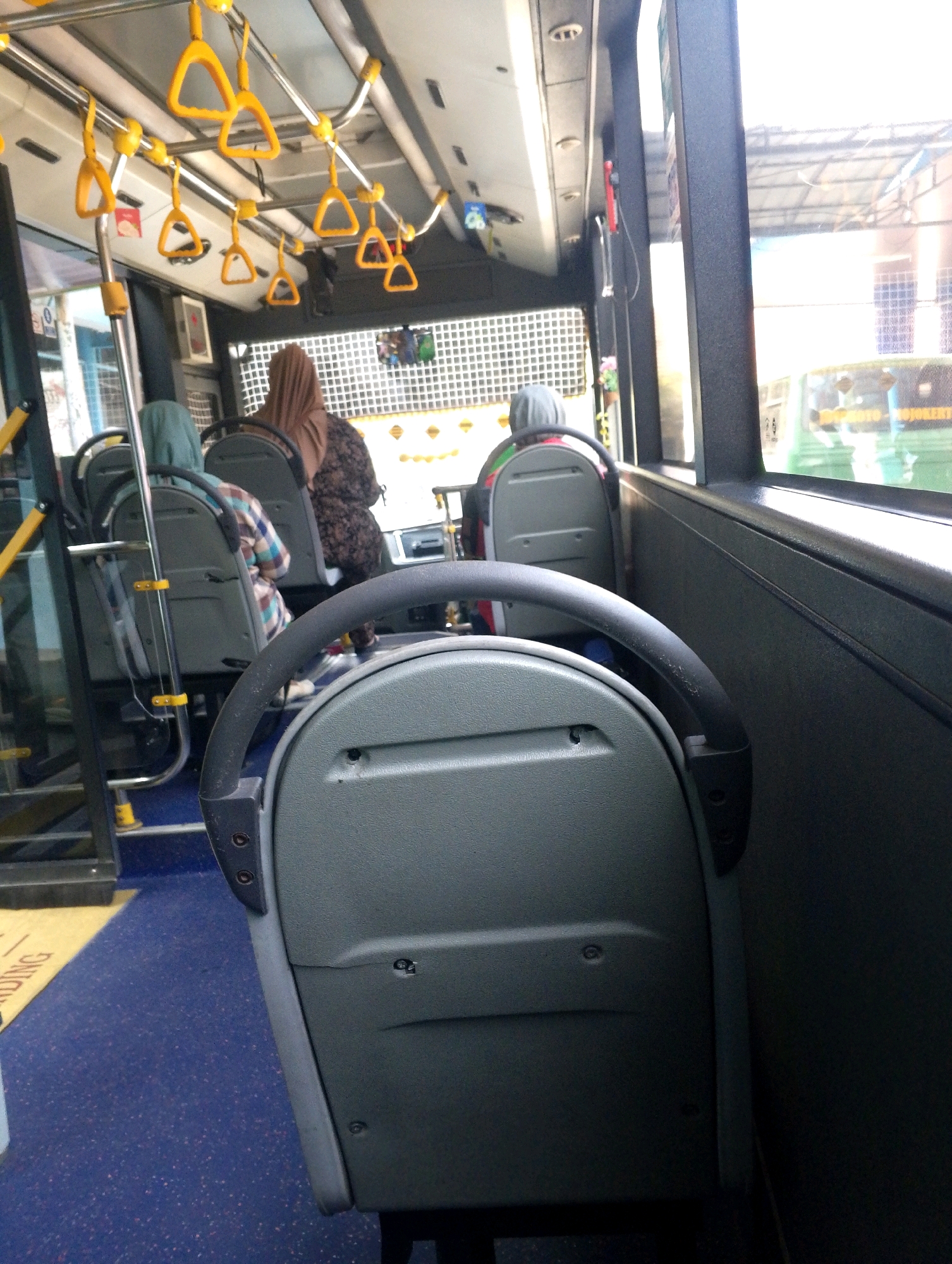 The image shows the interior of a bus. In the foreground, a gray plastic bus seat with a black handle is visible. In the background, several other bus seats are occupied by passengers wearing headscarves. Yellow grab handles are attached to the ceiling of the bus. Outside the bus window, a green bus is visible, along with a building structure. The floor of the bus is blue with flecks of color, and there is a yellow mat with text on it.