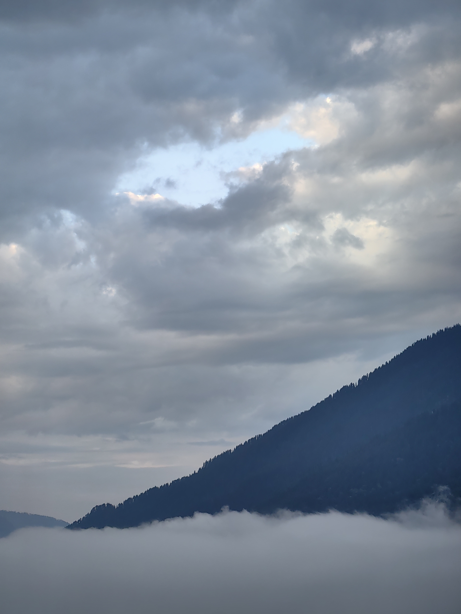 A wide-angle, vertical shot captures a mountainous landscape partially obscured by clouds. The top portion of the image is dominated by a cloudy sky, with patches of lighter blue and hints of sunlight breaking through. The clouds are a mix of dark and light grays. The lower half of the image features the dark silhouette of a tree-covered mountain slope, rising diagonally from the bottom left to the top right. Below the mountain slope, a dense layer of white mist or fog blankets the lower visible terrain, creating a stark contrast with the dark mountain above. The image appears to be a natural landscape, likely Vashisht, India, given the reference, with no people, man-made structures, or specific actions visible. The overall mood is serene and atmospheric, with the interplay of light and shadow adding depth to the scene.