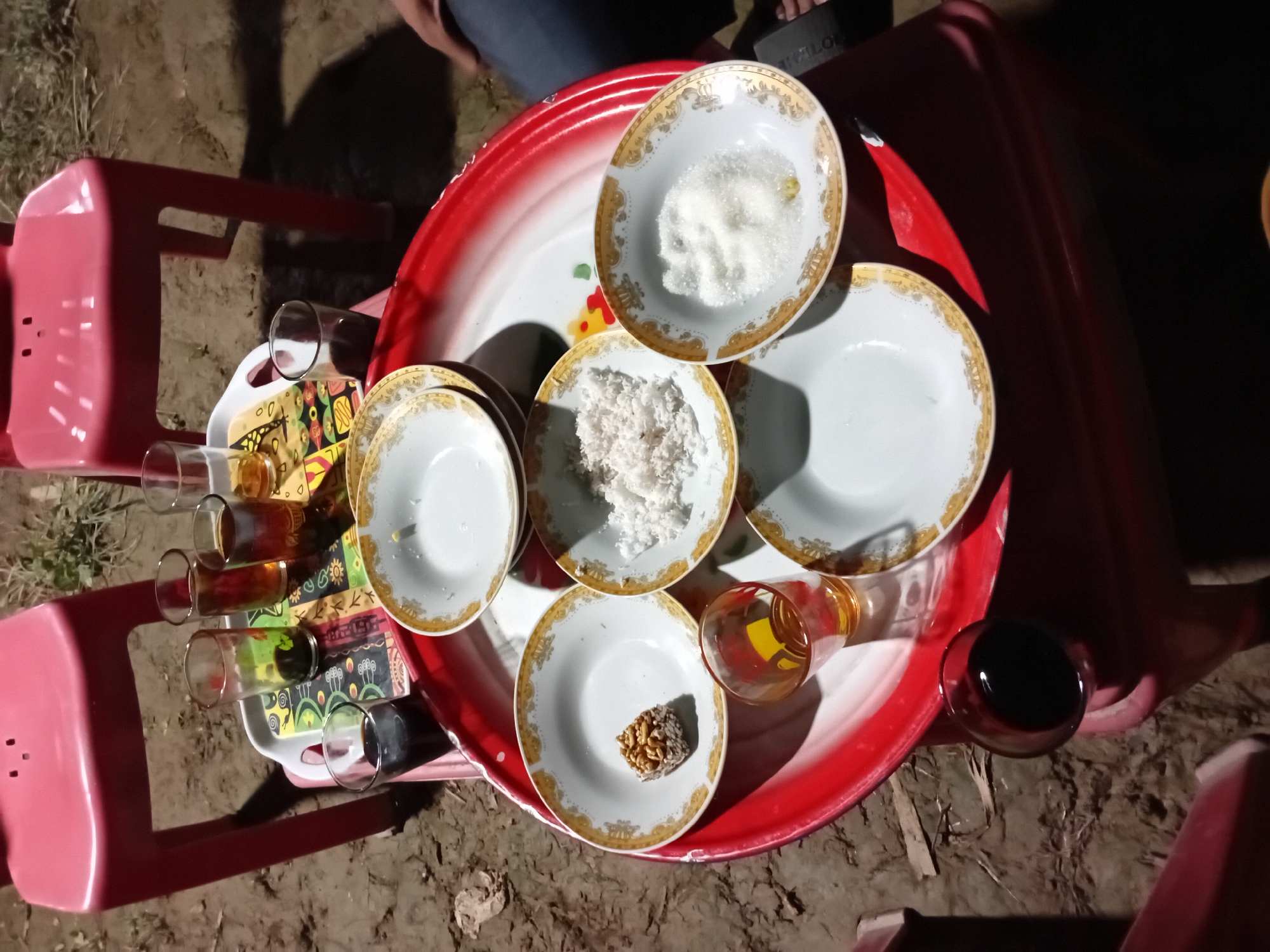 A high-angle shot captures a collection of dishes and drinks arranged on a red and white tray, which is placed on a red stool. The tray holds several small white plates with ornate gold trim. One plate contains white granular sugar, another has shredded white coconut, and a third holds a single, small, brown, square-shaped treat. Two clear glasses, one with amber liquid and the other with a dark liquid, are also on the tray, along with a small glass containing a golden liquid, likely a beverage.
To the left of the tray, another red stool is visible, supporting a tray with four clear glasses filled with amber-colored liquid. The arrangement suggests a serving of beverages and snacks. The scene is set outdoors on a dirt ground with some dry grass and leaves scattered around. The lighting indicates it is likely nighttime or very early morning, as there are strong shadows and the overall illumination is dim. A person's legs and lap, clad in blue jeans, are partially visible in the upper part of the image, suggesting they are seated nearby and possibly involved in the activity. The context provided (Aluebuya, Indonesia) hints at a cultural or social gathering. The mood appears casual and informal. There is no visible text in the image.