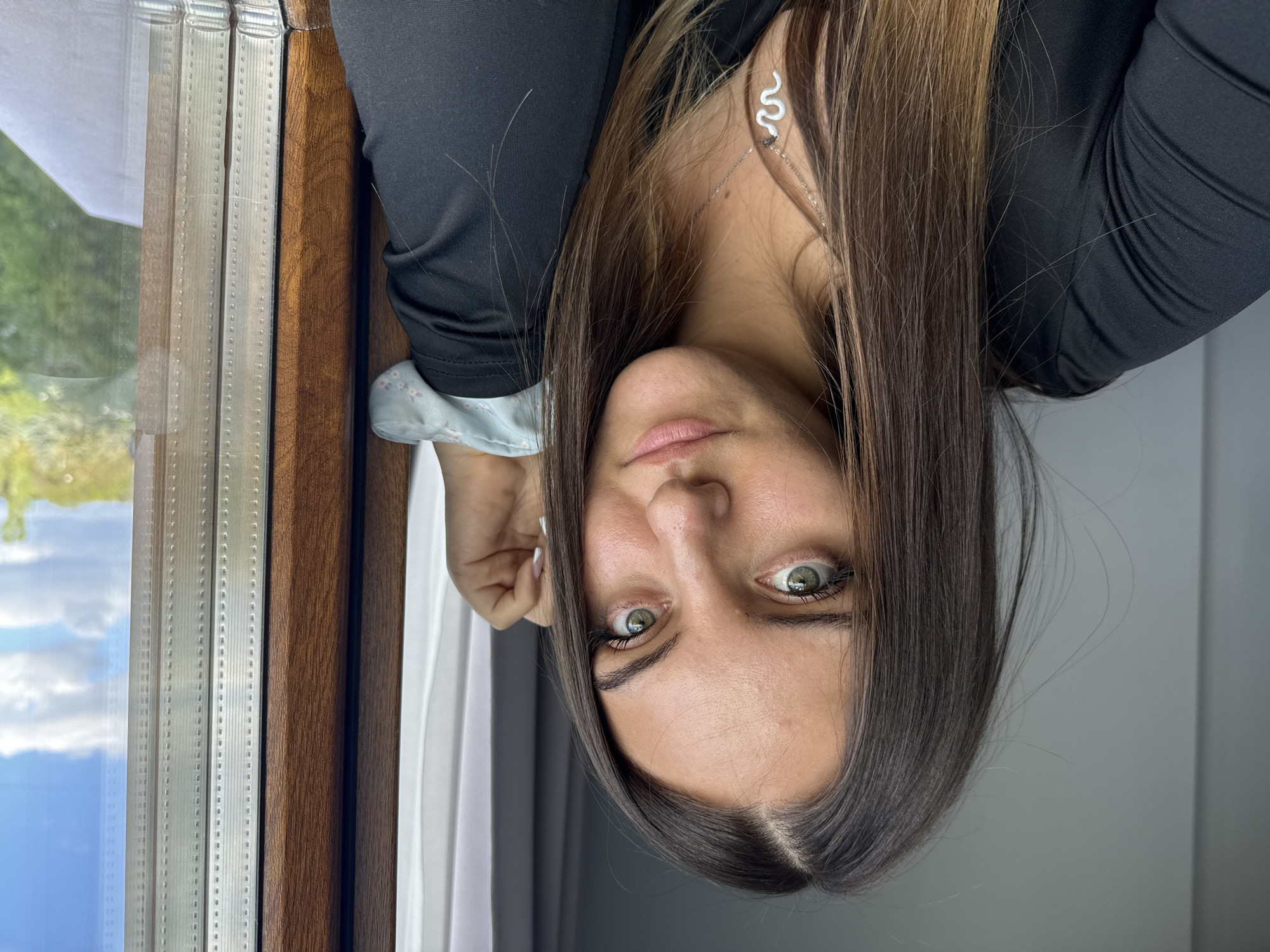 A young woman is visible in the image. She has long, dark hair with highlights, fair skin, and green eyes. She is wearing a black long-sleeved shirt and a silver snake-shaped earring and a necklace. She is partially leaning against a window frame that has multiple tracks with a wooden exterior. The window looks out onto a green landscape with a blue sky. There are sheer, light-colored curtains partially visible near the window. The location and city of Sofia, Bulgaria, cannot be determined from the image.