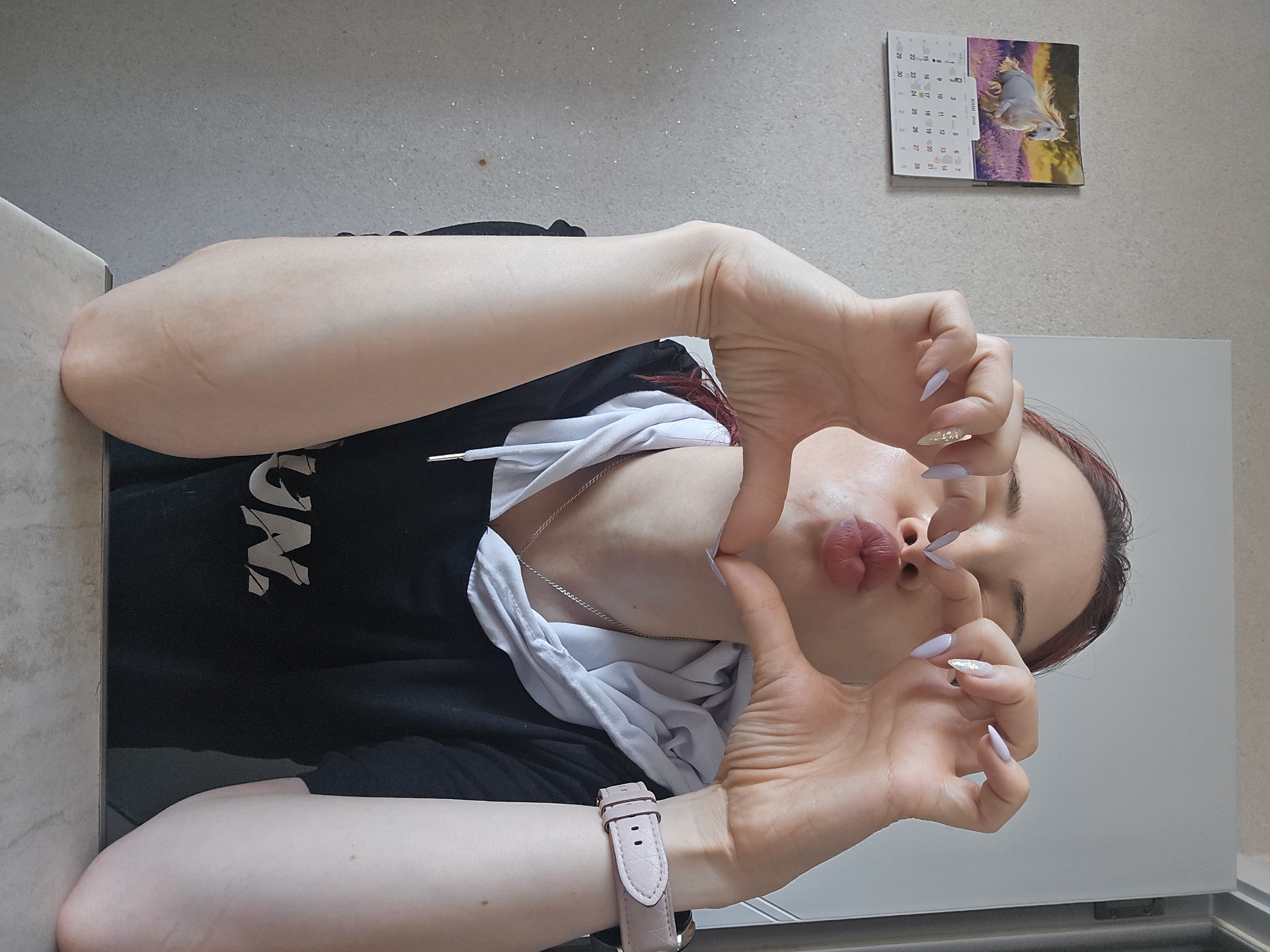 The image captures a young adult indoors, making a playful pose. She has long, reddish-brown hair and is wearing a black hoodie with a white drawstring and a silver chain necklace. Her nails are long, light purple, with some glitter detailing. A pink-strapped watch is visible on her left wrist. She is seen puckering her lips and forming a heart shape with her hands around her face.
The setting features light-colored walls or surfaces in the background and foreground, with a countertop visible on the left. On the wall behind her head, a small calendar is affixed. The calendar prominently displays "ЮНИ 2016" (June 2016) and features an image of a white horse in a field of lavender. Partial text reading "UN" is visible on her black hoodie. The scene takes place in Burgas, Bulgaria, as indicated by the context. The time appears to be daytime, or the room is well-lit by artificial light.