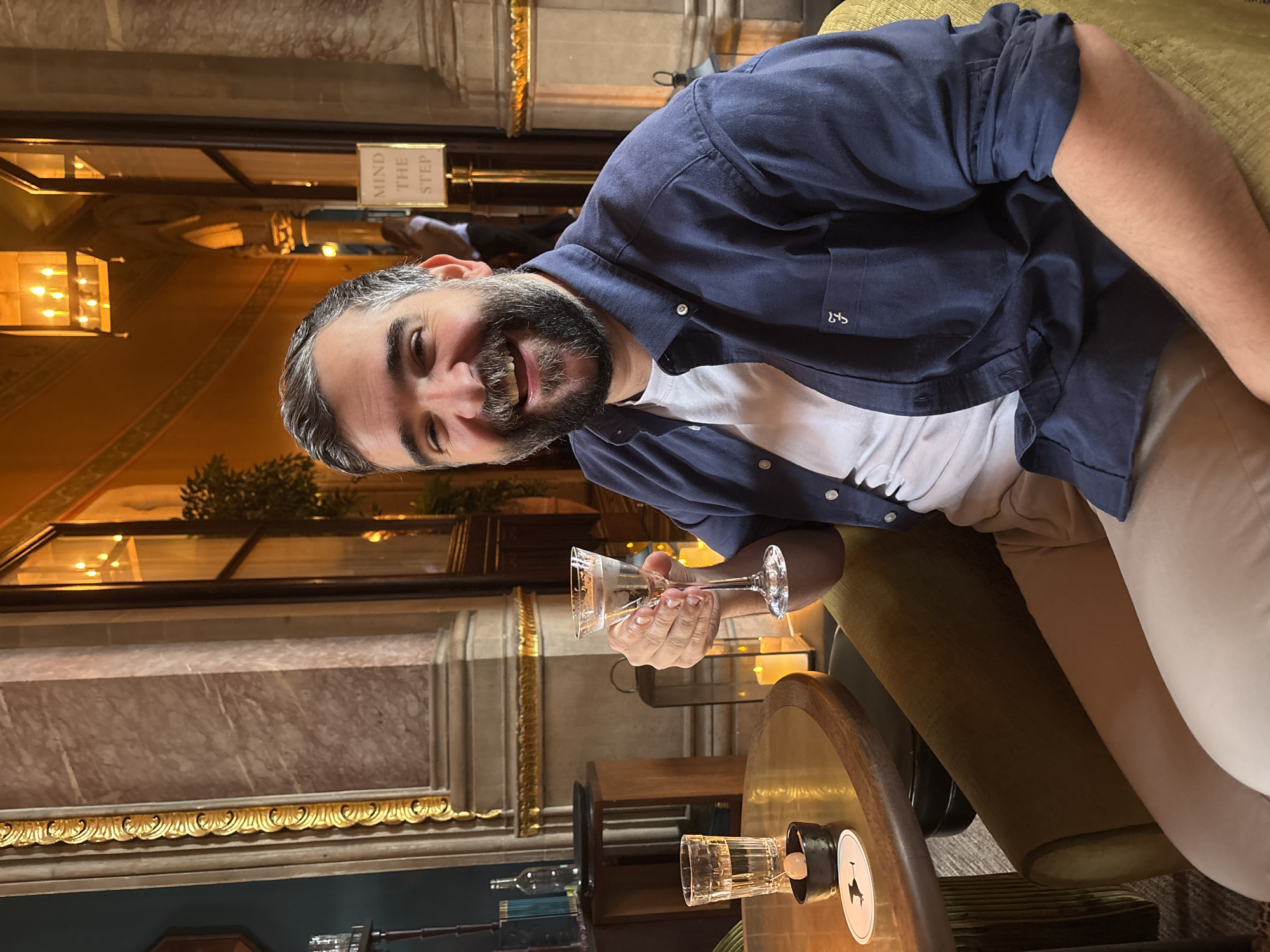 A man with a beard, wearing a navy blue shirt over a white t-shirt and tan pants, sits in a plush green chair at a small round table. He holds a stemmed cocktail glass, and another glass with a coaster sits on the table. The background features ornate architectural details, including marble columns, gold trim, and chandeliers. A sign reading "MIND THE STEP" is visible in the background. The location is likely a high-end bar in London, United Kingdom.