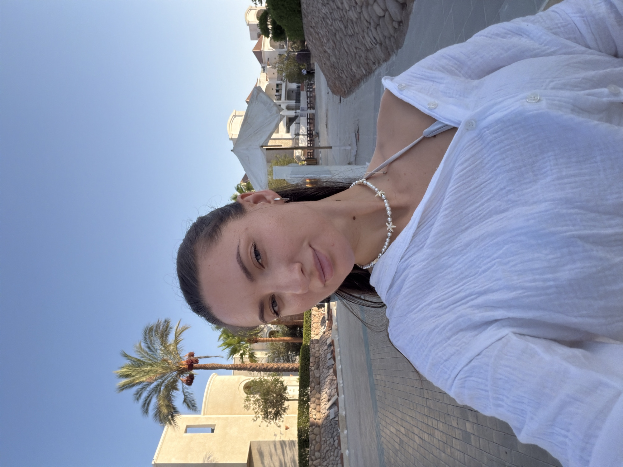 A woman is taking a selfie outdoors in Sharm El Sheikh, Egypt. She is wearing a white, open collared shirt over a light gray bikini top and a pearl necklace with shell-shaped pendants. Her dark hair is pulled back. Behind her, a paved walkway runs alongside a rocky embankment and a landscaped area with palm trees and buildings in the background. The sky is clear and blue.
