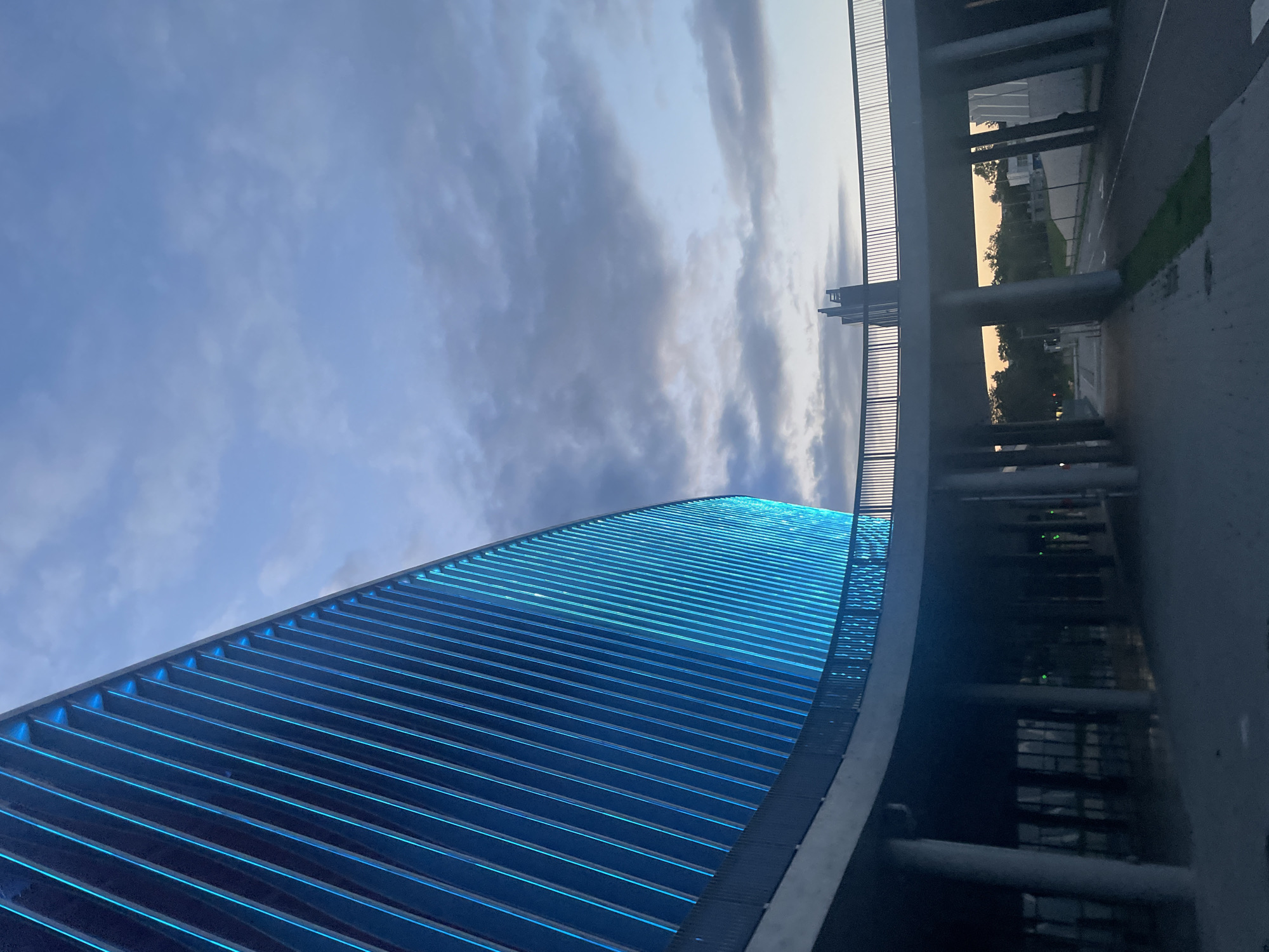 An exterior, low-angle, medium shot shows a modern building with a curved facade illuminated by blue vertical lights. The building is partially obscured by a curved elevated walkway with a metal railing. The walkway is supported by large concrete pillars. In the background, the sky is a gradient of light blue and gray clouds, with hints of orange near the horizon. A few trees and a road are visible beneath the walkway. The scene appears to be captured at dusk or dawn.