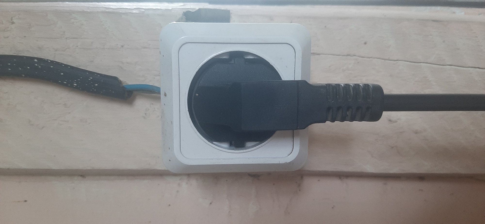A black electrical plug is inserted into a white wall socket. The socket is square with rounded corners and has a circular opening. Above the socket, a black electrical cord with white speckles is taped to the wall, with a blue wire visible near the socket. The wall appears to be textured and painted a light color, with some scuff marks and a vertical crack. The image is a close-up, centered on the electrical outlet and plug. The location is identified as Yessentukskaya, Russia.