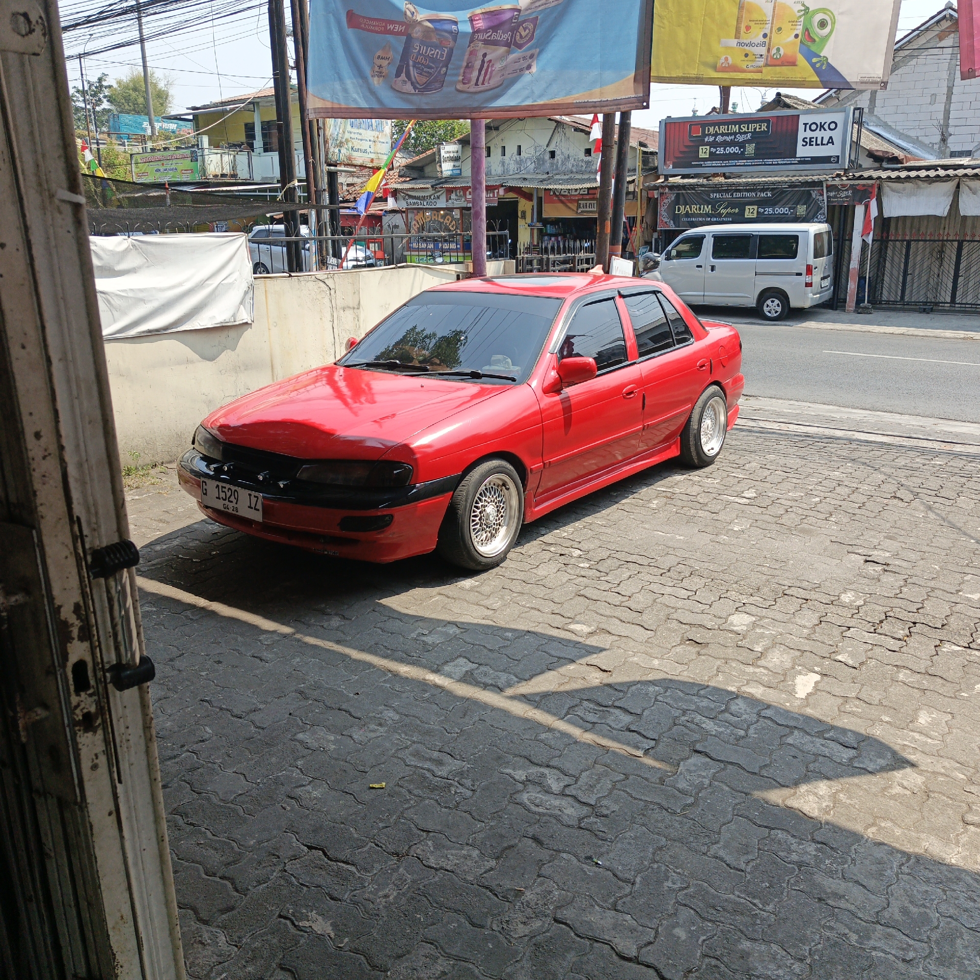 In Semarang, Indonesia, a bright red sedan with custom silver rims is parked on a cobblestone street. The car, a Kia Sephia, has tinted windows and a license plate that reads "G 1529 IZ". In the background, a white van is parked, and several shops with awnings are visible. Banners displaying advertisements for various products, including diapers and cigarettes, hang above the shops. The sky is clear, and the sun casts shadows on the street. The scene appears to be a typical day in a bustling urban area.