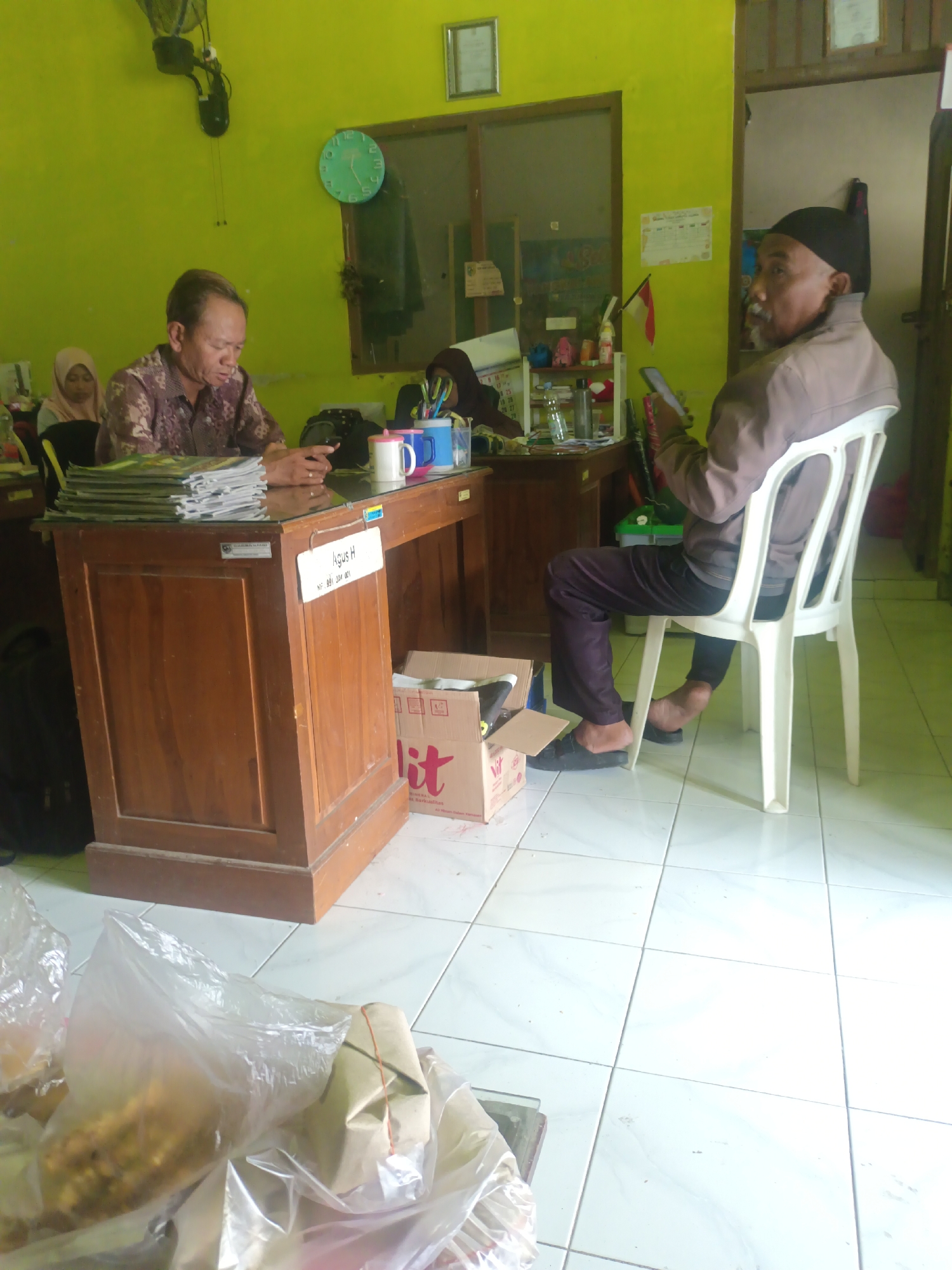 The image depicts an indoor scene that appears to be an office or administrative area in Sarimulyo, Indonesia. Two men are visible, along with two women partially obscured in the background.
In the foreground, a man with short, dark hair and a patterned brown shirt is seated behind a wooden desk, looking down at his phone. A stack of magazines or papers is to his left. On the desk, there are two mugs, one pink and one blue, and a pen holder with writing utensils. A sign on the desk identifies him as "Agus H".
To the right, an older man with a black cap and a light brown jacket is seated on a white plastic chair, also looking at his phone. He is facing away from the camera, with his legs crossed and bare feet visible.
The room has lime green walls. A large analog clock with a green face is mounted on the wall. A wooden cabinet is to the left of the desk. In the background, a door is visible, and a shelf with various items, including bottles and what appears to be a small Indonesian flag, is mounted on the wall. A cardboard box with red lettering is on the floor to the right of the desk. In the lower portion of the image, several plastic bags containing food items are on the floor. The floor is tiled with white and light gray squares.