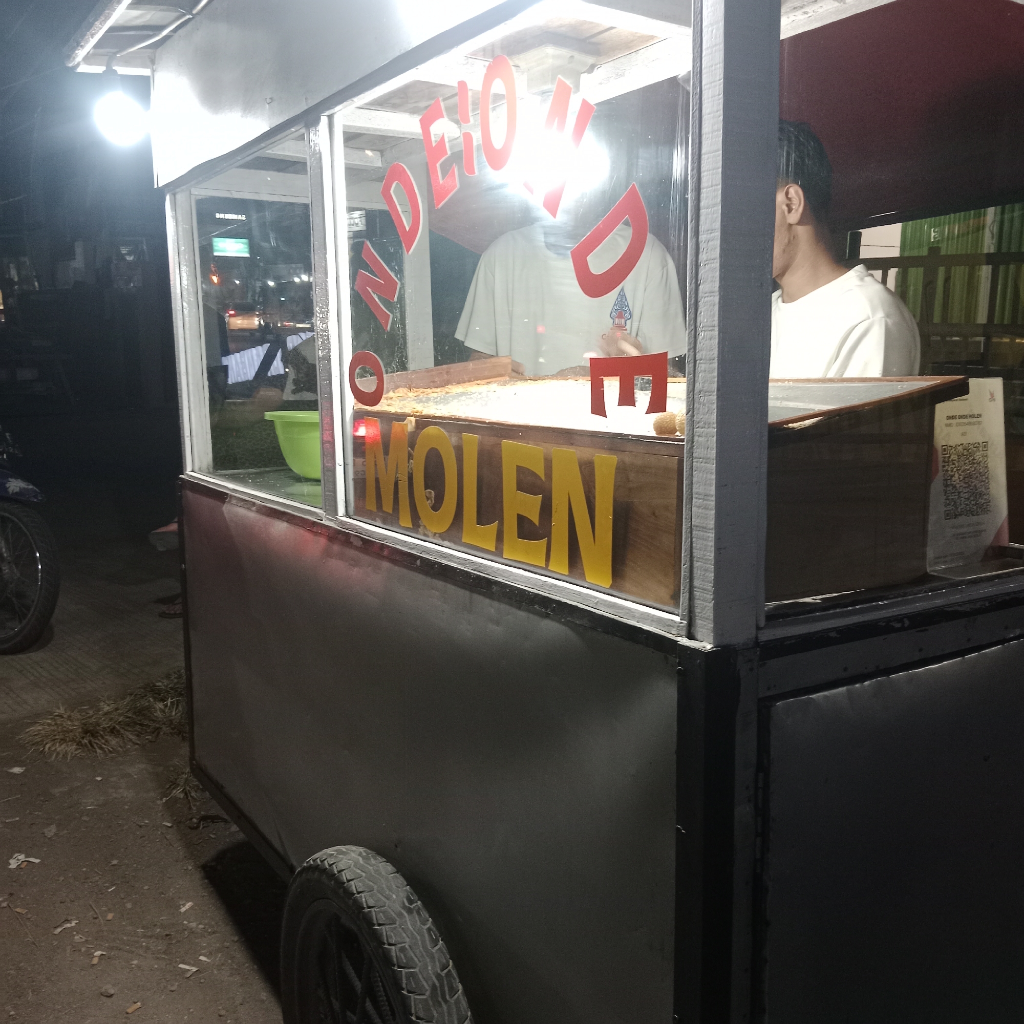 This is a photograph of a mobile food cart at night, specifically located in the city of Karangawen, Indonesia. The cart is illuminated by artificial lighting, casting a warm glow on the scene. The front of the cart features a glass display window where the words "NONDE MOLEN" are prominently displayed in red and yellow lettering. Behind the glass, a person wearing a light-colored t-shirt is partially visible, appearing to be preparing food. A green bowl and various food items are also visible within the cart. To the right of the cart, a QR code is displayed on a stand, likely for payment or information. The ground surrounding the cart is paved and appears to be dusty, with some scattered debris, including cigarette butts and dry grass. A portion of a motorcycle wheel is visible on the left side of the frame, suggesting its proximity to the cart. The overall environment suggests a street-side vendor operation in an urban setting.