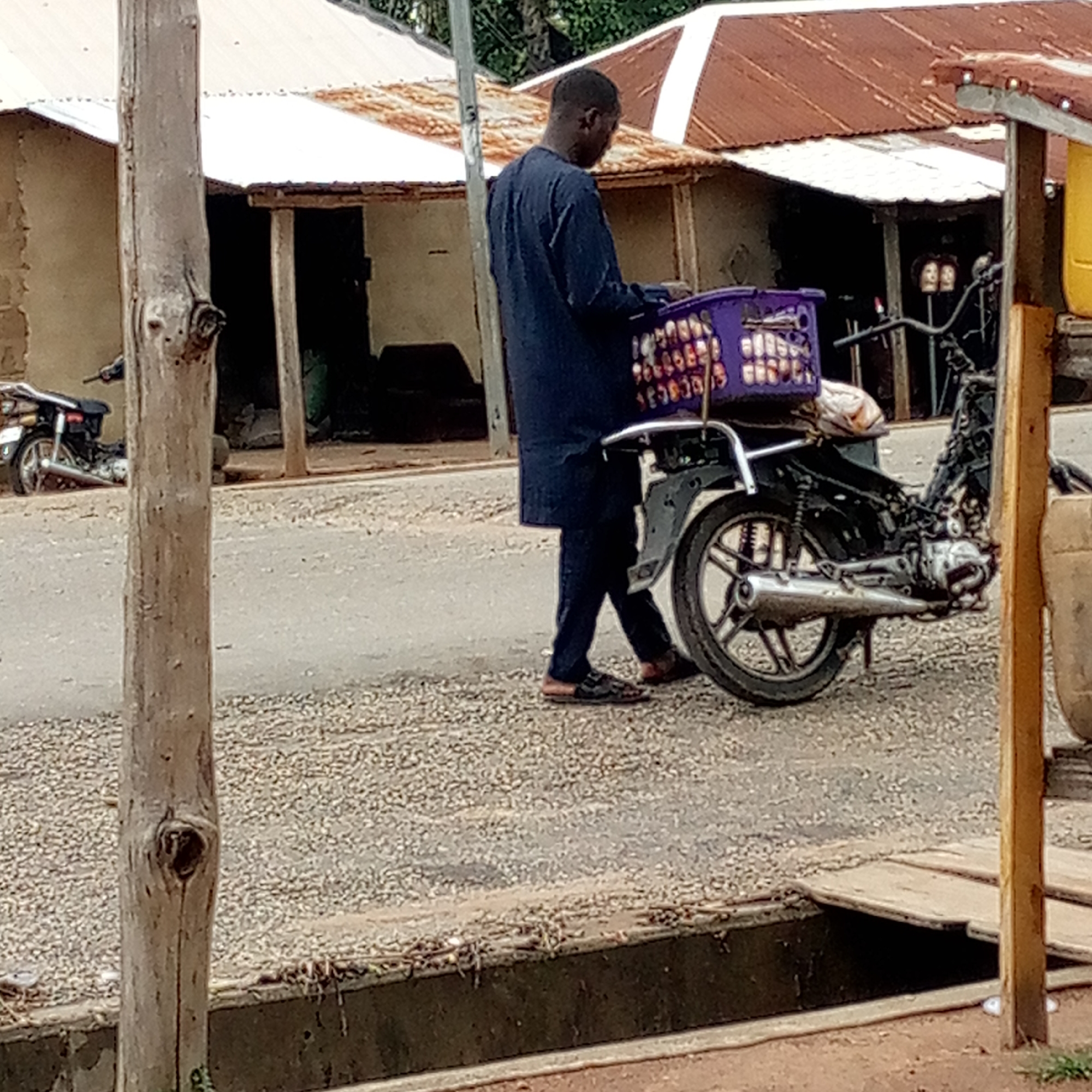 The photo shows a man standing next to a parked motorcycle on a paved road in Mararaba, Nigeria. The man is wearing a dark long-sleeved shirt and dark pants. On the motorcycle's rear carrier is a purple basket filled with items. In the background, there are buildings with corrugated metal roofs. A wooden post is visible in the foreground. The road appears to be lined with gravel. A drainage ditch runs along the edge of the road. Another motorcycle is parked further down the street.