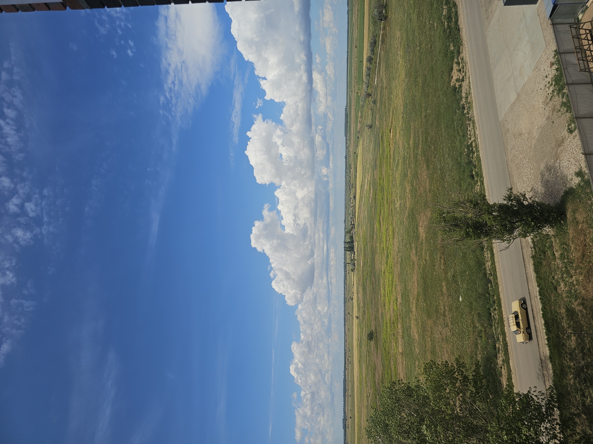 A landscape view captures a vast, flat, grassy field under a bright blue sky filled with scattered white clouds. A paved road runs horizontally across the middle ground, with a light-colored vintage sedan driving on it. Trees are visible on the left and right sides of the road, with more sparse vegetation and distant structures dotting the horizon. A fence separates a gravel and concrete area in the foreground from the field. The scene appears to be in the city of Skadovsk, Ukraine.