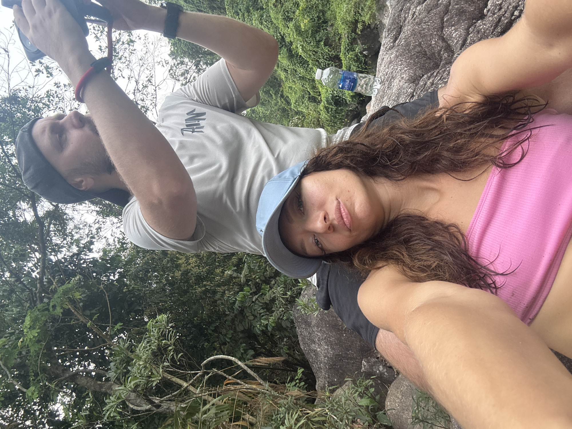 The image is a selfie taken outdoors in a tropical or subtropical environment. A man and woman are positioned close together. The man is slightly behind the woman, holding a camera up and away from himself, presumably to take the picture. He is wearing a grey t-shirt and a dark-colored cap. The woman is in front, wearing a pink top and a light blue cap. Her hair is wavy and dark.
A clear plastic water bottle is visible on a rock behind the man. The background consists of lush green foliage, indicating a natural setting. A portion of a rock formation is visible, where they seem to be sitting.
Given the information available, I can not positively identify the location in the city of Đà Nẵng, Vietnam.
