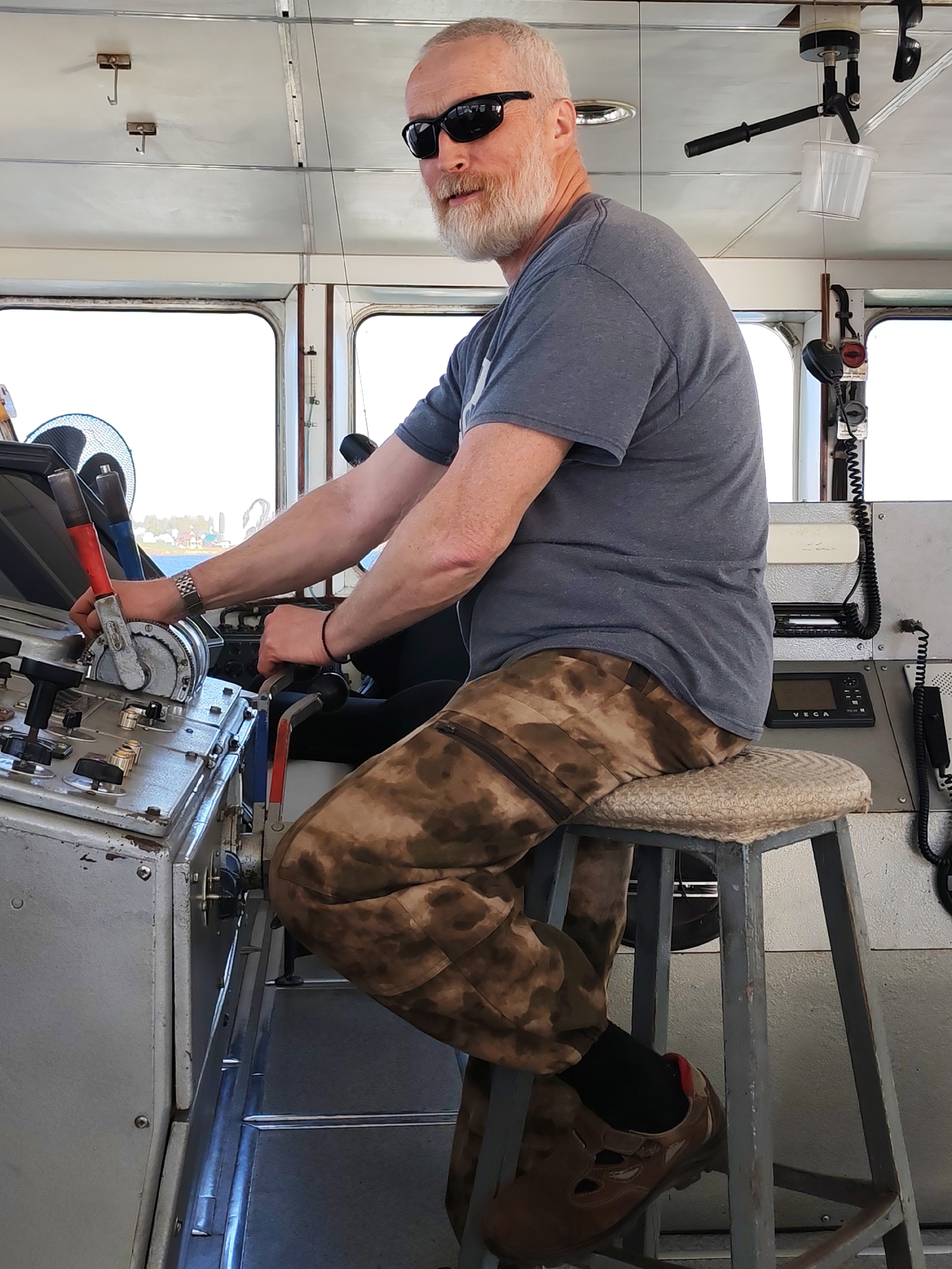 A gray-haired man with a beard and sunglasses sits on a stool in what appears to be the cockpit of a ship or boat. He wears a gray t-shirt and camouflage pants, with brown shoes visible on his feet. He is positioned at the controls, with his hands on levers and switches, suggesting he is operating the vessel. The cockpit is predominantly gray, with various control panels and equipment visible. Through the windows, a view of land and water is discernible. The scene appears to be indoors.