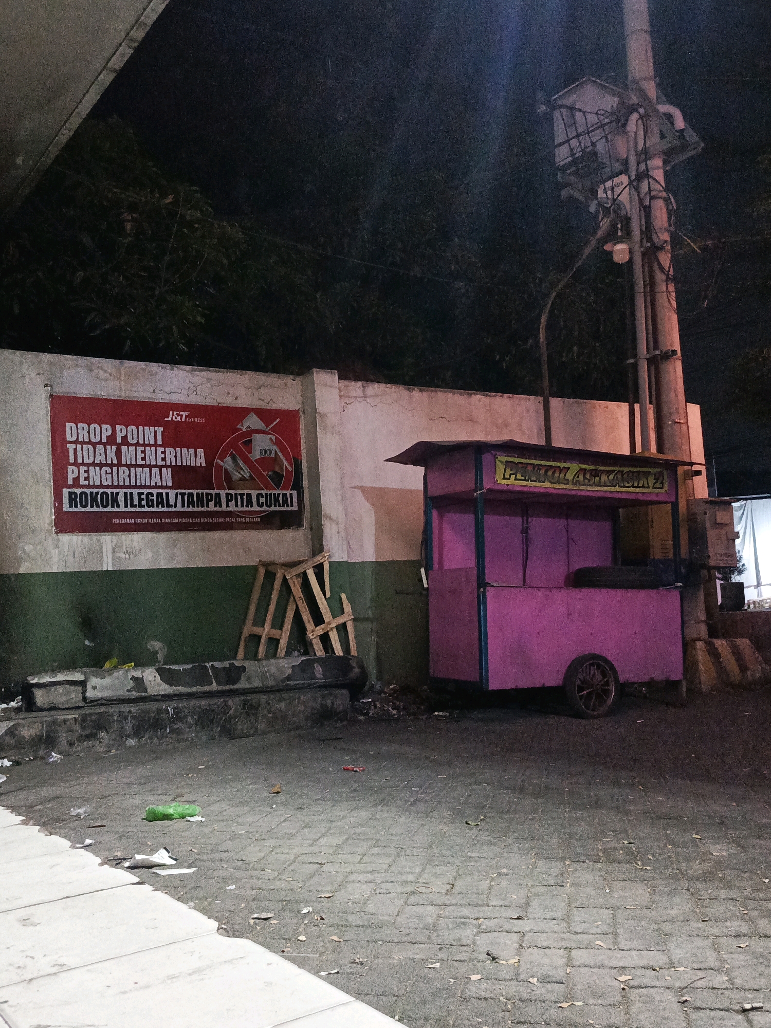 This is a medium-angle, exterior shot of a street scene in Pamekasan, Indonesia, at night. The image captures a pink food cart labeled "PENTOL AS KASIK 2" with a yellow sign. To the left of the cart is a concrete wall with a red banner that reads "DROP POINT TIDAK MENERIMA PENGIRIMAN ROKOK ILEGAL/TANPA PITA CUKAI," which translates to "Drop Point does not accept illegal cigarette shipments/without excise tape." The wall is partially covered by a green-painted section. In front of the wall, there are some discarded concrete blocks and wooden scaffolding. The ground is paved with cobblestones and shows scattered debris, including a green plastic bag and cigarette butts. A utility pole with electrical wires and a transformer box stands to the right of the food cart. The background is dark and features foliage, indicating the presence of trees. The lighting is dim, suggesting it is nighttime.