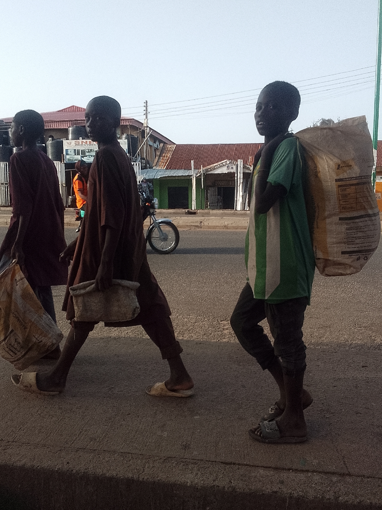 Three young individuals are walking along the sidewalk of a street in Pantame, Nigeria. They appear to be in transit, as evidenced by the large sacks and containers they are carrying. The first individual on the left is wearing a maroon outfit and carries a bag. The second is wearing a brown tunic and carrying a small square bag. The third and rightmost individual is wearing a green and white striped t-shirt and black pants, carrying a large sack over their shoulder. They are walking toward the camera. In the background, there's a motorcycle parked on the road, buildings with a red roof, and a clear sky. The photo has a somewhat muted color palette, suggesting it was taken in the early morning or late afternoon.