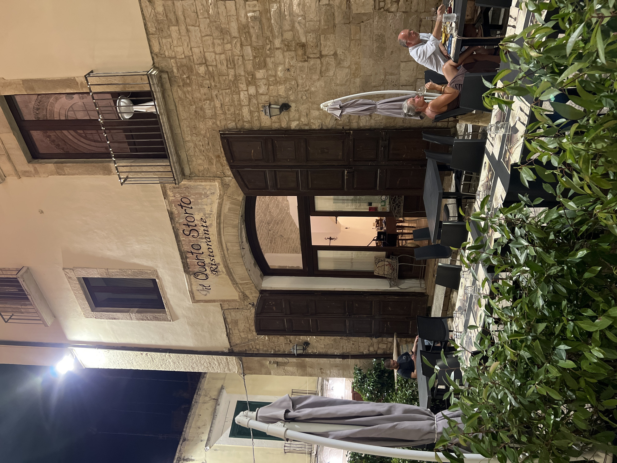 The image shows the exterior of a restaurant in Bitonto, Italy, at night. The restaurant's name, "il Quarto Storie Ristorante," is visible above the entrance. There are two tables set up outside the restaurant with chairs and place settings.
In the foreground, lush green foliage partially obscures the view of the tables. On the left side of the image, a closed umbrella stands next to one of the tables. A man is seated at this table, looking at his phone.
Further to the right, two people are seated at another table. A woman with gray hair, wearing a dark dress, is holding a glass of white wine. A man with glasses, wearing a light blue shirt, is also seated at this table.
The restaurant building is constructed of stone with cream-colored stucco. Above the entrance are large wooden double doors that are partially open, revealing a dimly lit interior. A balcony with a metal railing is present on the upper floor of the building, and a light fixture is visible on the balcony. A street lamp illuminates the scene from the left. The overall atmosphere is that of a quiet evening at an Italian restaurant.