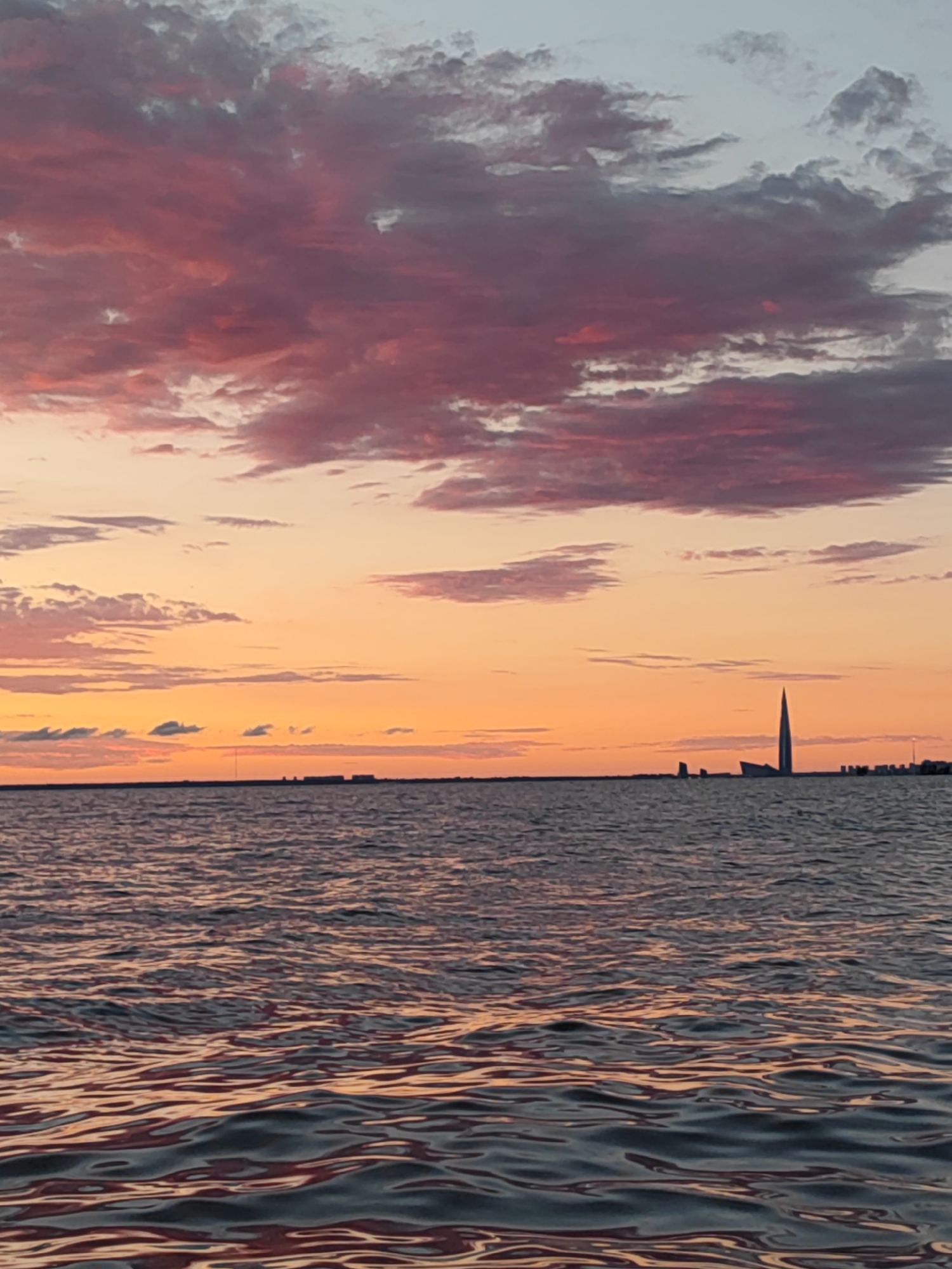The image captures a sunset over the Gulf of Finland in Saint Petersburg, Russia. The sky is painted with vibrant hues of pink, orange, and light blue, with scattered clouds reflecting the fading sunlight. The calm waters of the gulf mirror the colors of the sky, creating a serene and picturesque scene.
In the distance, along the horizon, the iconic silhouette of the Lakhta Center, a modern skyscraper, is visible. The building's distinctive spired design stands out against the colorful sky. Other buildings and structures, typical of a city's waterfront skyline, are also discernible. The foreground is dominated by the rippling surface of the water, reflecting the warm tones of the sunset. There are no people or discernible actions in the image, creating a tranquil and contemplative atmosphere.