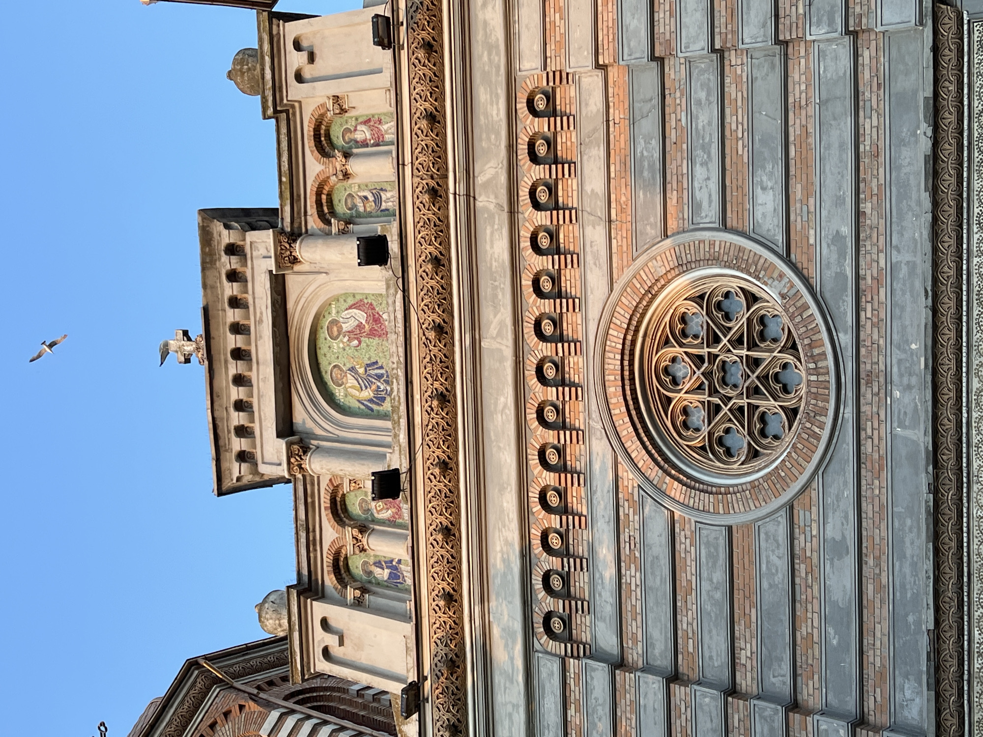 The image captures the facade of the Orthodox Cathedral of Saints Peter and Paul in Constanța, Romania, under a clear blue sky. A seagull is visible in flight against the sky, positioned in the upper portion of the frame. The cathedral's architecture features intricate details, including mosaics depicting religious figures above arched openings, ornate friezes, and a large circular window with Gothic tracery. The building exhibits a combination of brick and stone elements, with some signs of weathering and age. Lighting suggests it is either early morning or late afternoon, casting a warm glow on the facade.