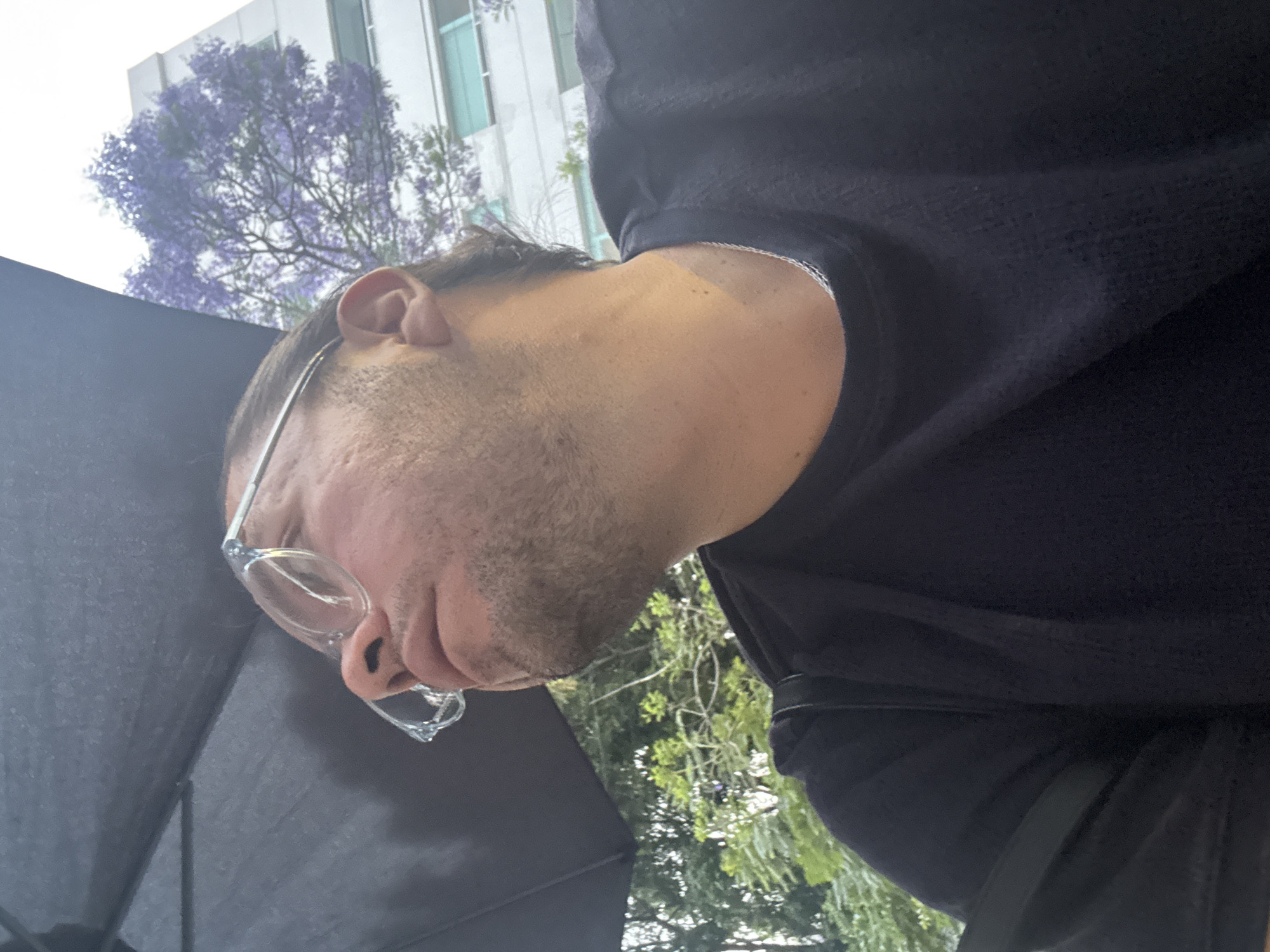 A tilted close-up shot depicts a person's face wearing glasses, situated under an umbrella. The background reveals a building and trees, including a Jacaranda tree identifiable by its purple flowers. The location is presumed to be Mexico City, Mexico, based on the presence of Jacaranda trees.