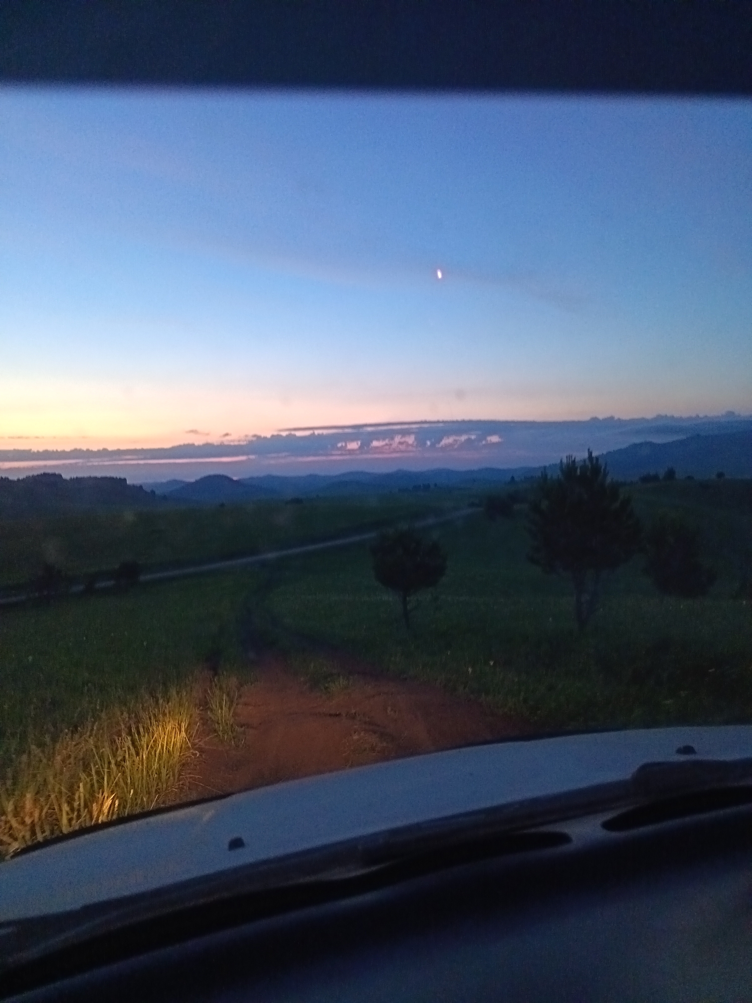 The image depicts a rural landscape at dusk, viewed from inside a vehicle. The sky is a gradient of deep blue to pale pink near the horizon, with a faint crescent moon visible. Rolling hills and distant mountains form the background, shrouded in twilight. In the mid-ground, a dirt road winds through a grassy field dotted with trees. The foreground is dominated by the interior of the car, specifically the dashboard and windshield, with the headlights illuminating the tall grass at the edge of the road. The overall impression is one of peaceful solitude in a natural setting. The location is identified as Sadovyi, Russia, implying a rural or semi-rural environment within that region.