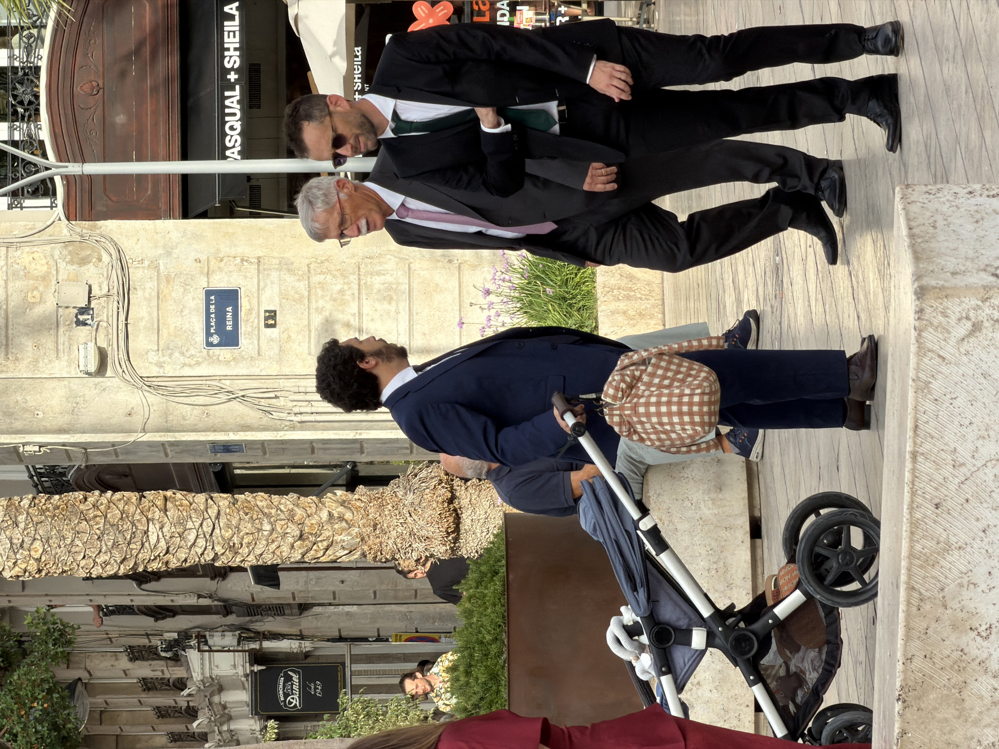 Three men in formal attire are walking on a paved street next to a palm tree and buildings. The man on the left is wearing a dark blue suit and a light blue shirt with a dark tie. He is holding the handle of a stroller. The middle man is wearing a dark gray suit, a white shirt, and a pink patterned tie. He has glasses and a mustache. The man on the right is wearing a black suit, a white shirt, and a green tie. He is wearing sunglasses. A stroller with a beige checkered bag on the handle is in the foreground. In the background, a building has a sign that says "PLAÇA DE LA REINA". Another building has a sign that says "PASQUAL + SHEILA". The scene is lit by daylight.