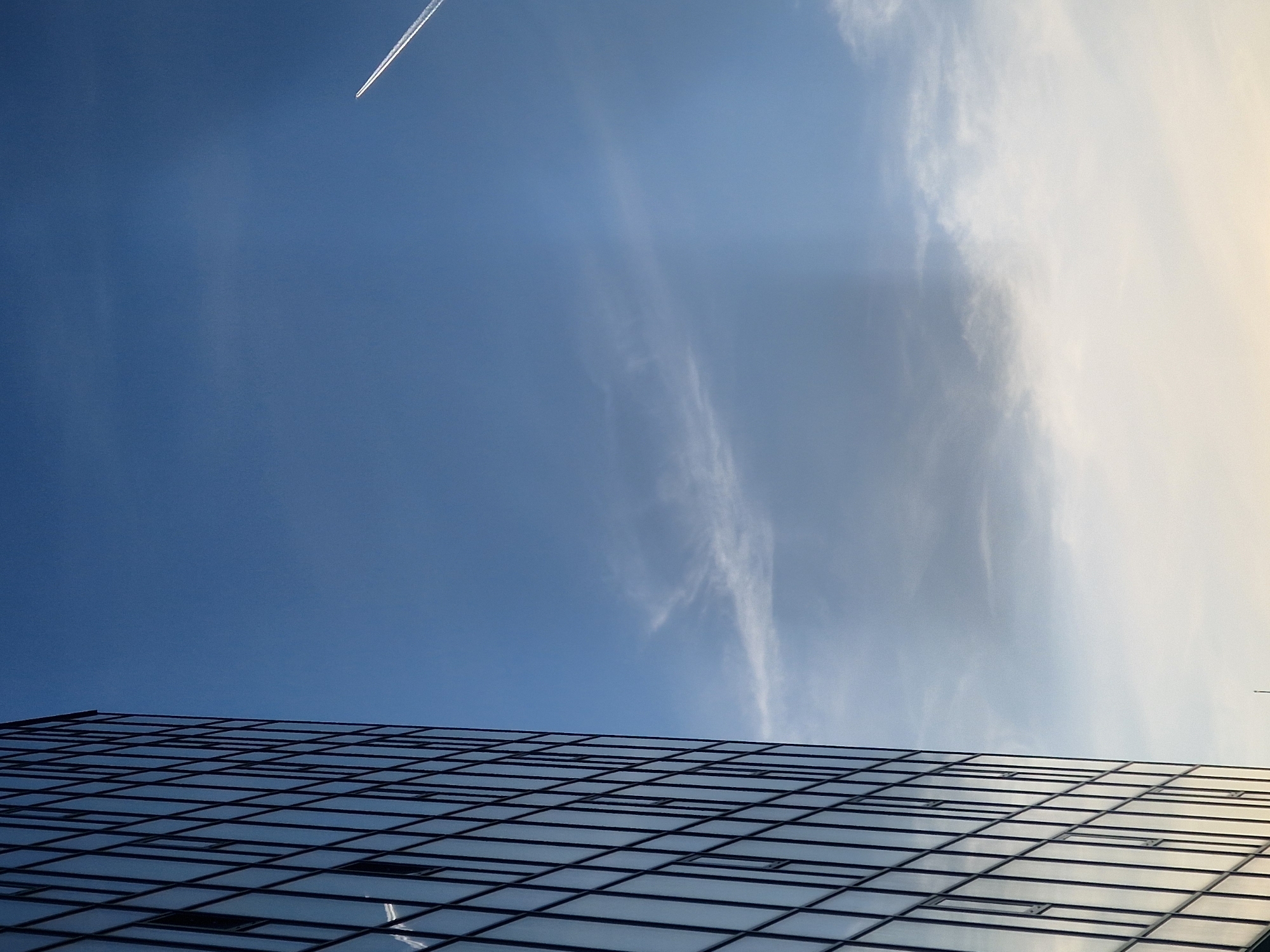 The image, when viewed upright, captures a clear blue sky above a modern glass building. The building occupies the lower portion of the frame, showcasing a grid pattern of windows that reflect the sky and light. Some windows appear to be slightly open or recessed.
The sky, filling the upper two-thirds of the image, transitions from a deep blue on the left to a lighter, brighter blue with a hint of yellowish white on the right, suggesting the direction of the sun. Wispy, white clouds are scattered across the sky, becoming denser towards the brighter right side. High in the upper left quadrant, a distinct, straight white contrail from an unseen airplane cuts across the blue expanse.
There is no specific activity depicted beyond the static presence of the building and the fading contrail of a distant aircraft. The time of day appears to be daytime, likely late morning or afternoon, given the bright illumination and the soft yellow hue in part of the sky. The weather is clear with scattered high-altitude clouds. No specific location cues are visible beyond the contemporary architecture. There is no visible text in the image.