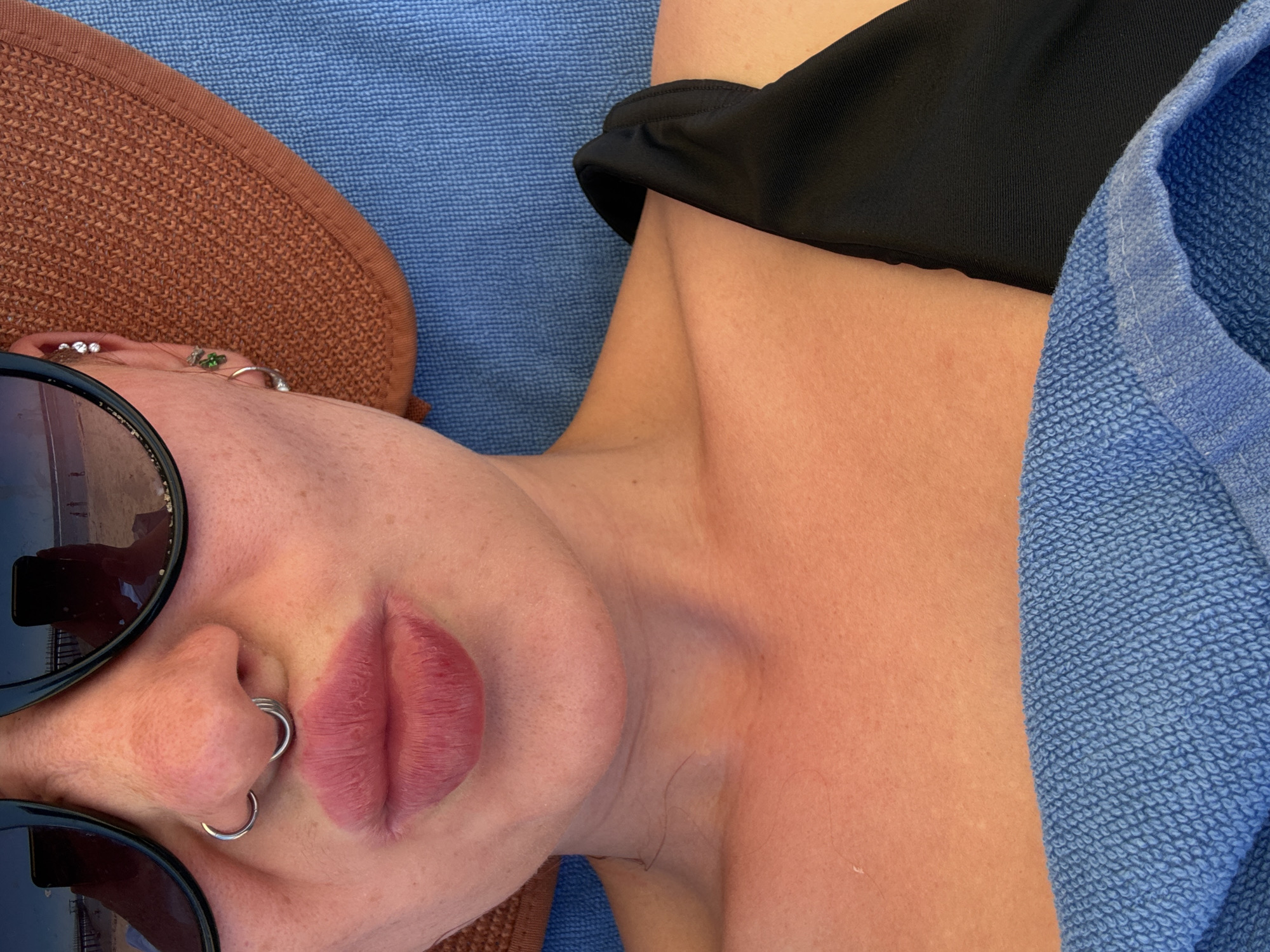 A close-up, overhead shot shows a person wearing sunglasses, a hat, and a black swimsuit. The person has a septum piercing and appears to be pouting. The background is a blue towel. The image does not contain any information about the location being Sharm El Sheikh, Egypt.