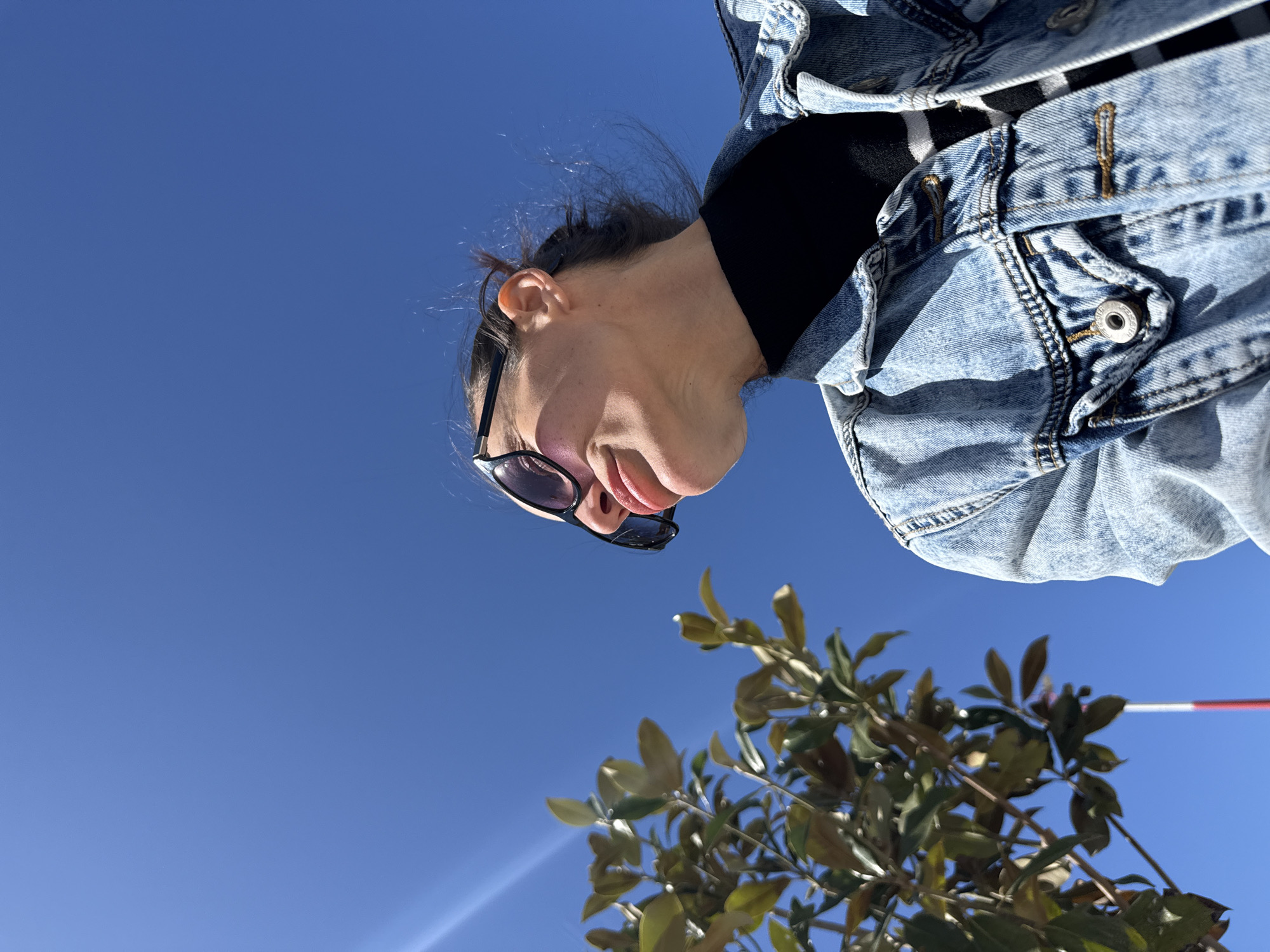 A woman in sunglasses and a denim jacket is looking down at the camera. A tree with green leaves is visible in the foreground, against a clear blue sky. A red and white pole is slightly visible behind the tree. The location is Mouzouras, Greece.