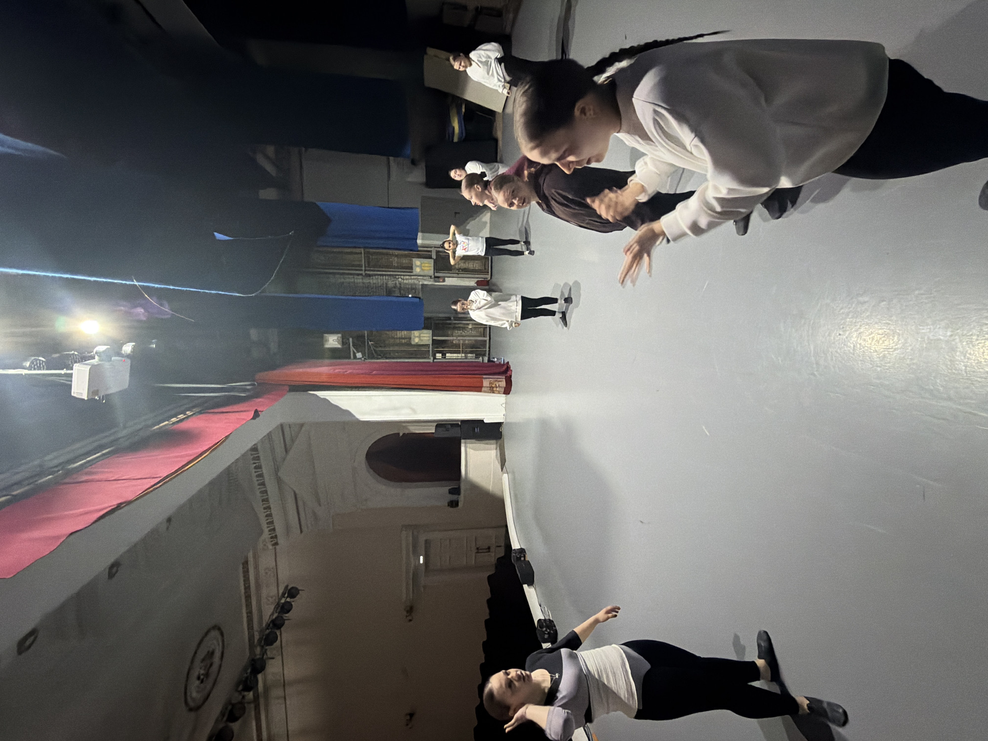 The image shows a group of dancers rehearsing in a theater in Perm, Russia. The dancers are aligned in a row on a stage and are performing some sort of choreographed movement, possibly involving their hands. The stage is bare, with a grey floor. A red curtain hangs on the side of the stage. There are several people on the stage, and they seem to be focused on rehearsing some sort of dance. There are theater seats in the background.