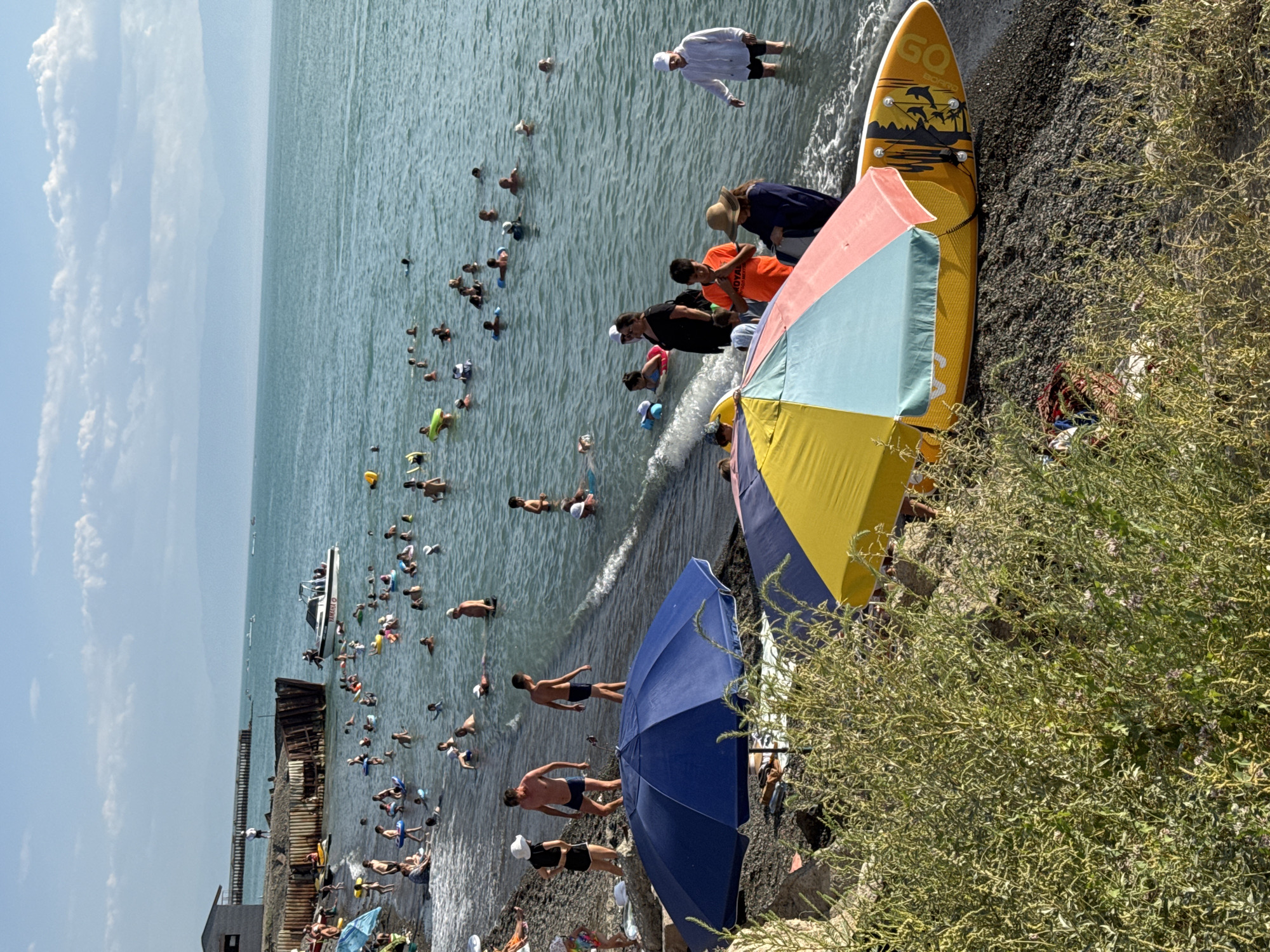 The image depicts a lively scene at a beach, likely in Zharbulak, Kazakhstan, on a clear day with a bright blue sky dotted with clouds. The turquoise water is calm, with small waves lapping at the shore, and populated by numerous individuals swimming and playing. A white motorboat with several occupants is visible in the water. The beach itself is dark gravel, with green vegetation growing along the edge. Two beach umbrellas, one blue and one colorful (yellow, blue, and pink), are set up on the shore. A yellow paddleboard with black graphics is lying on the sand, partially covered by one of the umbrellas. Several people are on the beach, some standing and some sitting. One person in a white jacket and dark shorts is standing in the shallow water, looking towards the shore. In the background, a long pier extends into the water, and distant mountains are visible on the horizon. The overall impression is one of a popular recreational spot enjoying warm weather.