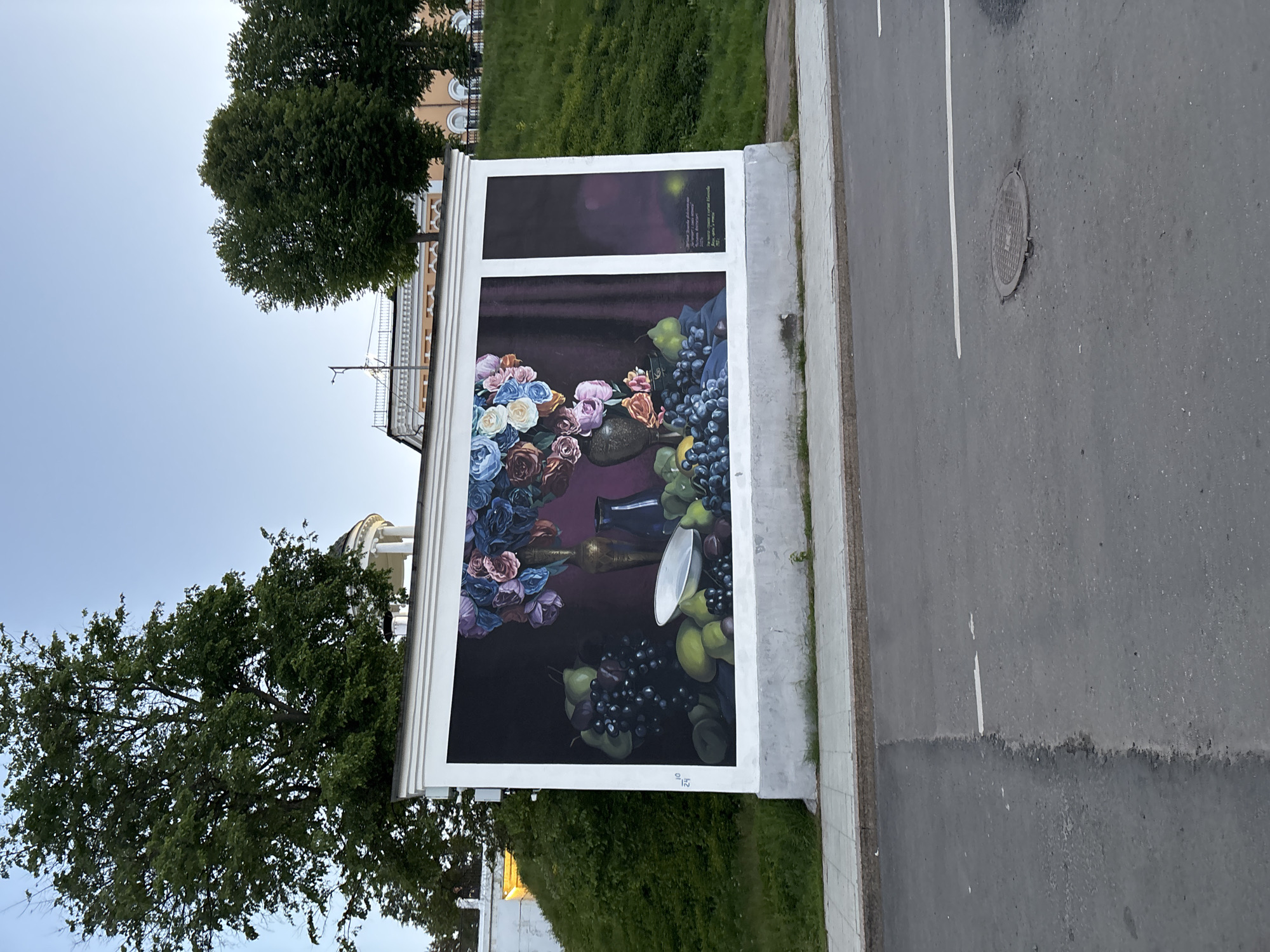A large outdoor art installation in Rusanovo, Russia, features a still life painting depicting flowers, fruit, and vases, with a dark purple background. The installation is mounted on a white frame or wall-like structure located beside a road. To the left of the installation are trees and a building with visible architectural details. To the right is a paved road with a manhole cover.
