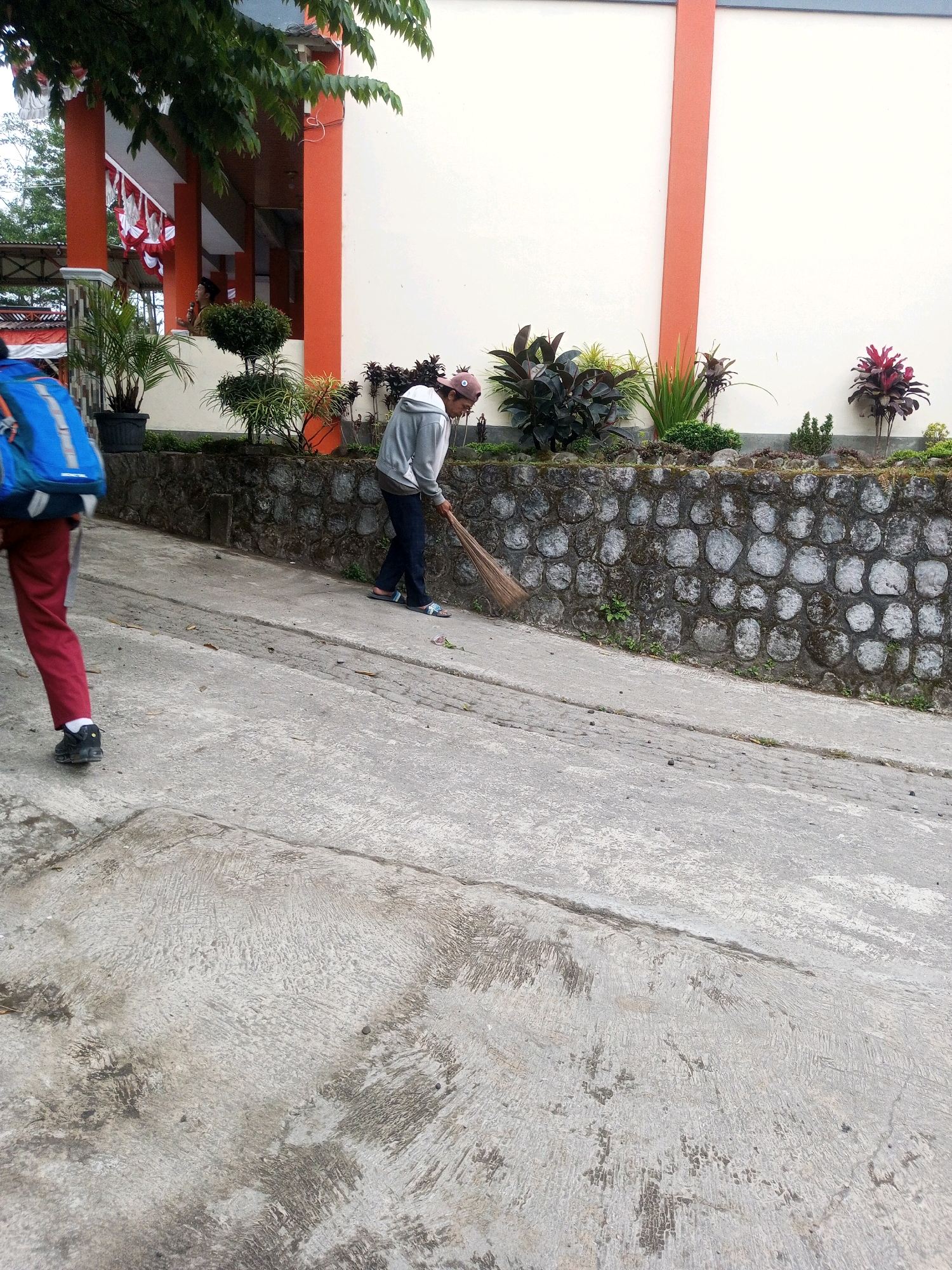 In Kalibeber, Indonesia, a person wearing a grey hooded sweatshirt, dark pants, and sandals is sweeping a concrete walkway with a broom. To the left, another person walks away from the camera, wearing a blue backpack and red pants. The walkway slopes downwards and appears to be outdoors, adjacent to a building with an orange trim. A stone wall separates the walkway from a garden area with various plants and some decorative elements. The building's facade is white with orange accents. The sky is overcast.