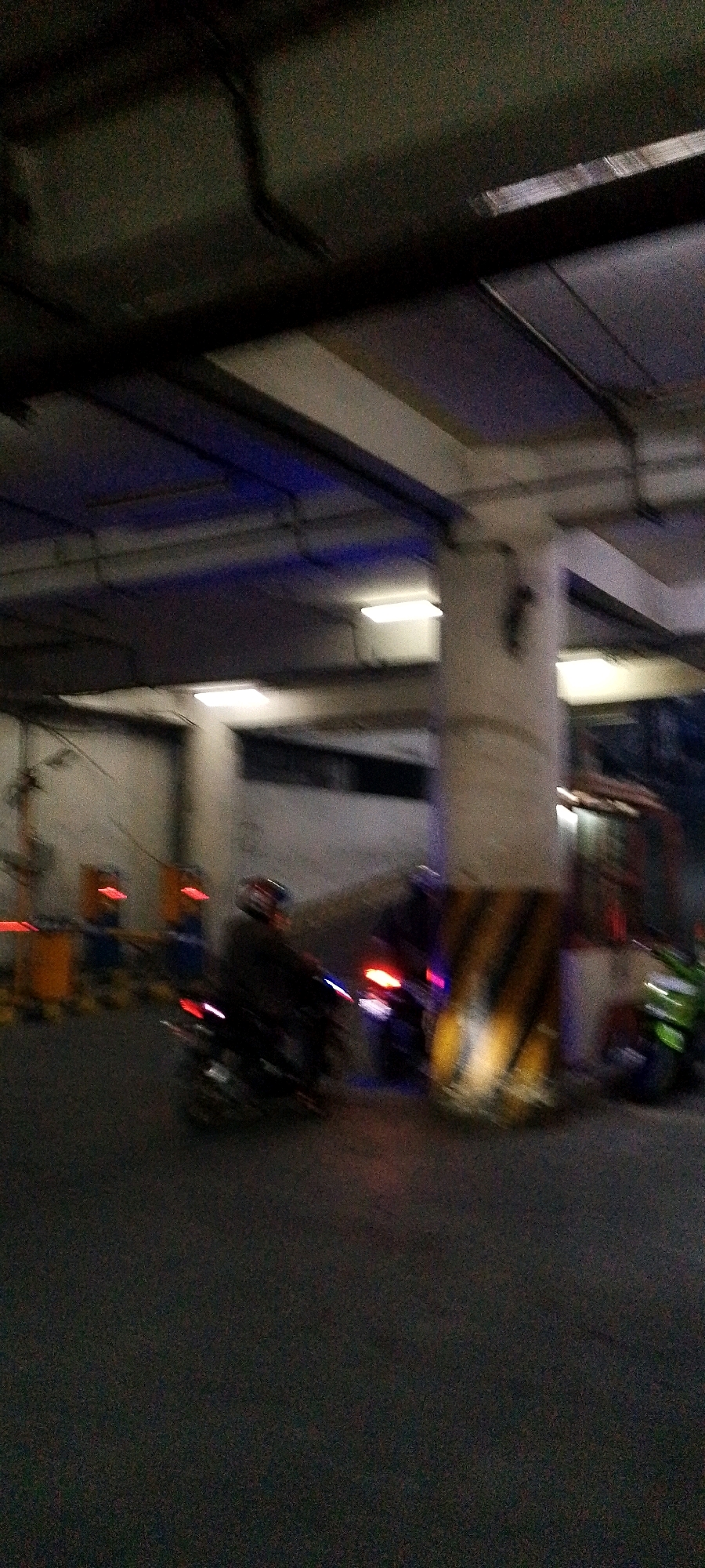 The image depicts a blurry, low-light scene, likely taken in an urban environment at night or in a dimly lit area. In the foreground, two motorcycles are visible, with one rider on each. The rider on the left is more prominently featured, wearing a helmet. The motorcycle on the left has its taillight illuminated. To the right of this motorcycle, another motorcycle is partially visible, also with its taillight lit.
In the mid-ground, several yellow and blue barriers with red lights are present, suggesting a controlled or restricted area, possibly an entrance or exit point. A large concrete pillar with black and yellow hazard stripes is also visible, supporting an overhead structure. Further back, a vehicle with a red roof is parked. The background is characterized by concrete walls and overhead structures, with some artificial lighting, including what appear to be rectangular fluorescent lights and some blue lighting. The overall impression is that of a parking garage or an industrial-type area.
The image quality is poor due to blur and low light, making precise identification of details difficult. There are no clear indicators in the image that definitively place the scene in Bandung City, Indonesia. However, based on the visual cues of motorcycles, traffic barriers, and urban infrastructure, it is plausible that this scene could be found in a city like Bandung, known for its traffic and infrastructure.