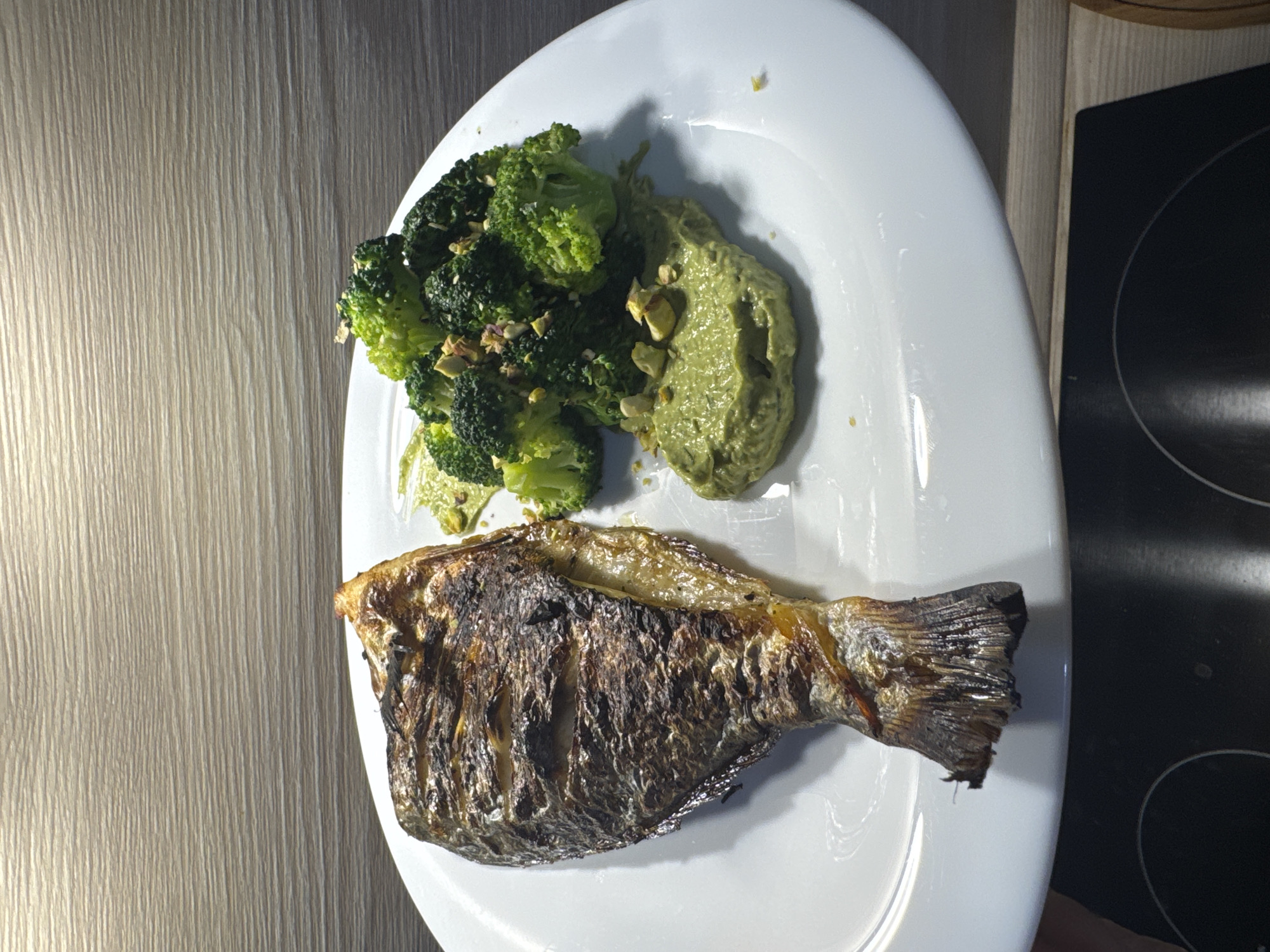 A whole grilled fish sits on a white plate next to a pile of steamed broccoli florets. The fish is golden brown with grill marks and appears to be seasoned. The broccoli is bright green and sprinkled with chopped nuts. A dollop of green sauce, possibly avocado or pesto, is smeared on the plate. The scene is set against a wooden-grained background. The image was taken indoors, likely in a kitchen, and there are no people visible.