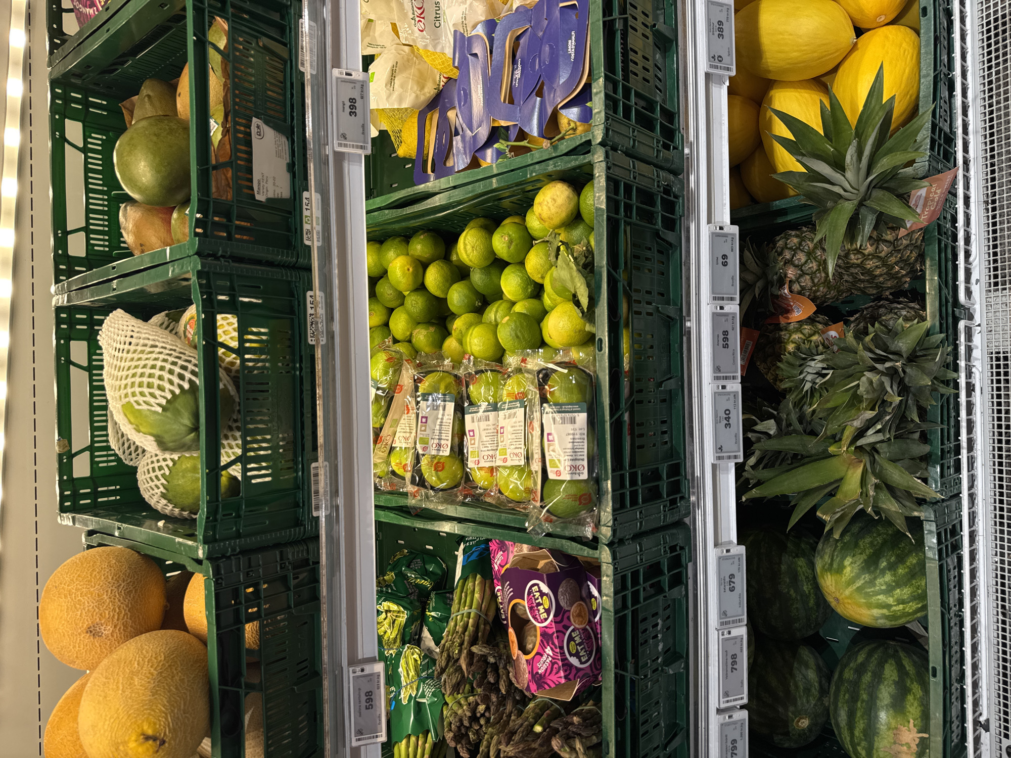 This image presents a close-up shot of produce stacked in green crates at a grocery store, likely located in Vik, Iceland. The produce is arranged vertically in the crates, including mangoes, cantaloupes, limes, lemons, pineapples, watermelons, asparagus, and passion fruits. Price tags are attached to the side of each column of produce. The crates are arranged to maximize the space.