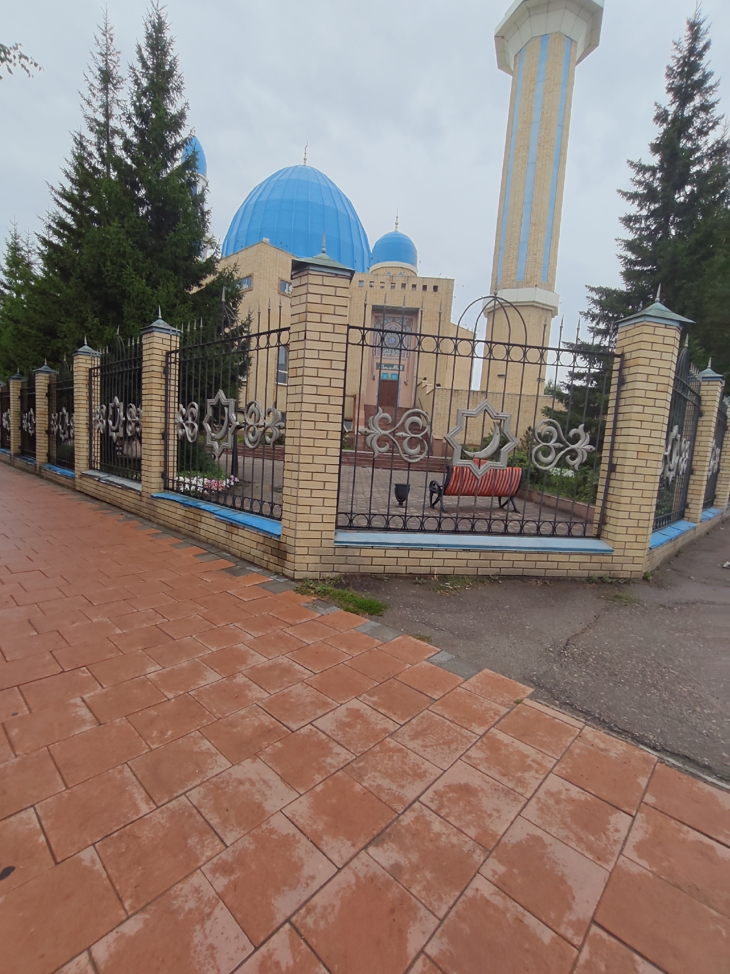 The image captures a view of a mosque in Petropavl, Kazakhstan. A paved walkway in the foreground leads to a decorative metal fence with brick pillars. Beyond the fence, the mosque features a prominent blue dome and a tall, striped minaret. Evergreen trees surround the building. The sky is overcast, suggesting cloudy weather. There are no people visible in the image.