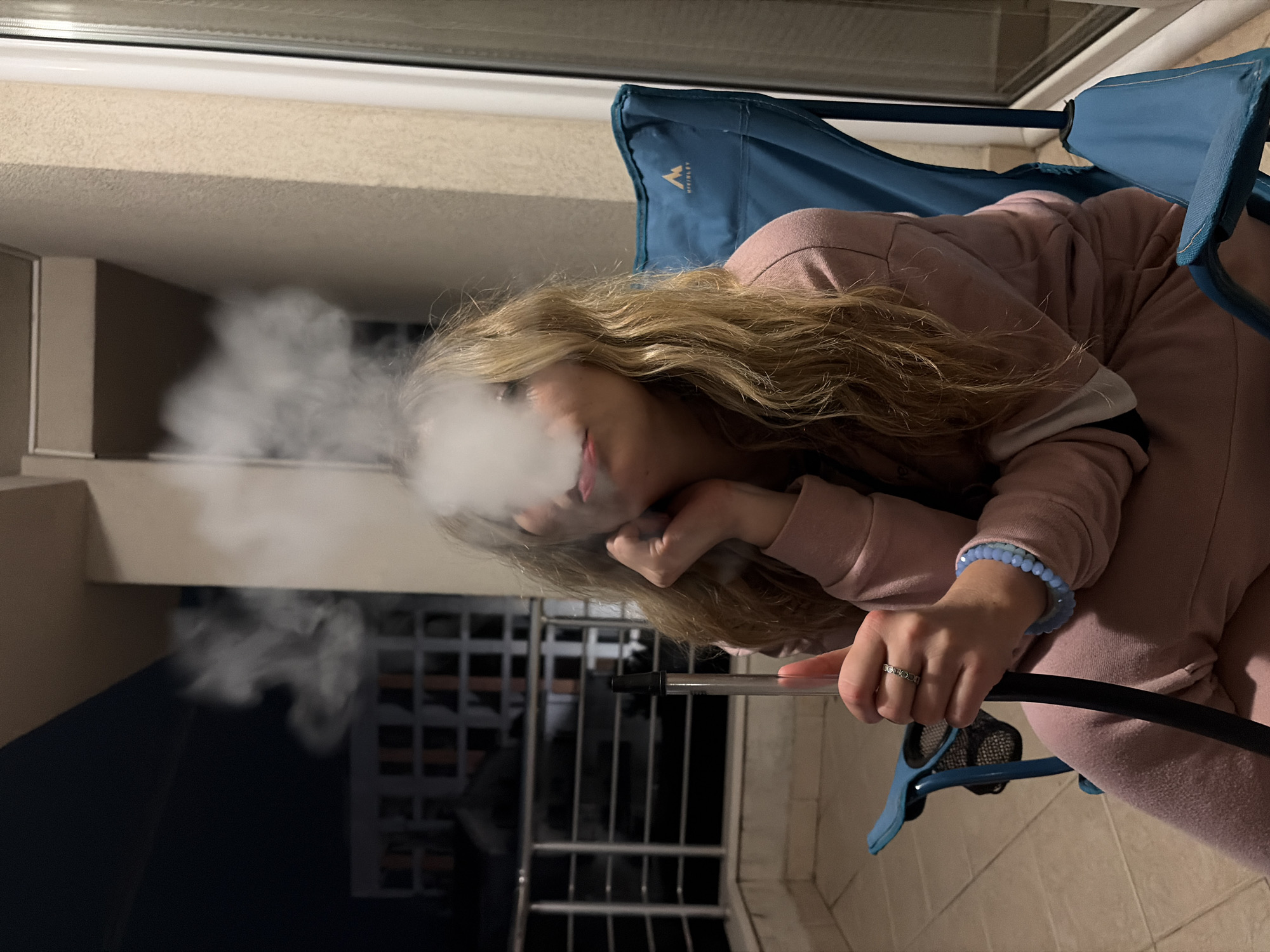A woman with blonde hair sits on a blue folding chair on a balcony. She wears a light pink tracksuit and a blue beaded bracelet. She holds a black hose and exhales smoke from her mouth. The balcony has a tiled floor and a metal railing. In the background, there are buildings and a dark sky. The location is Budva, Montenegro.