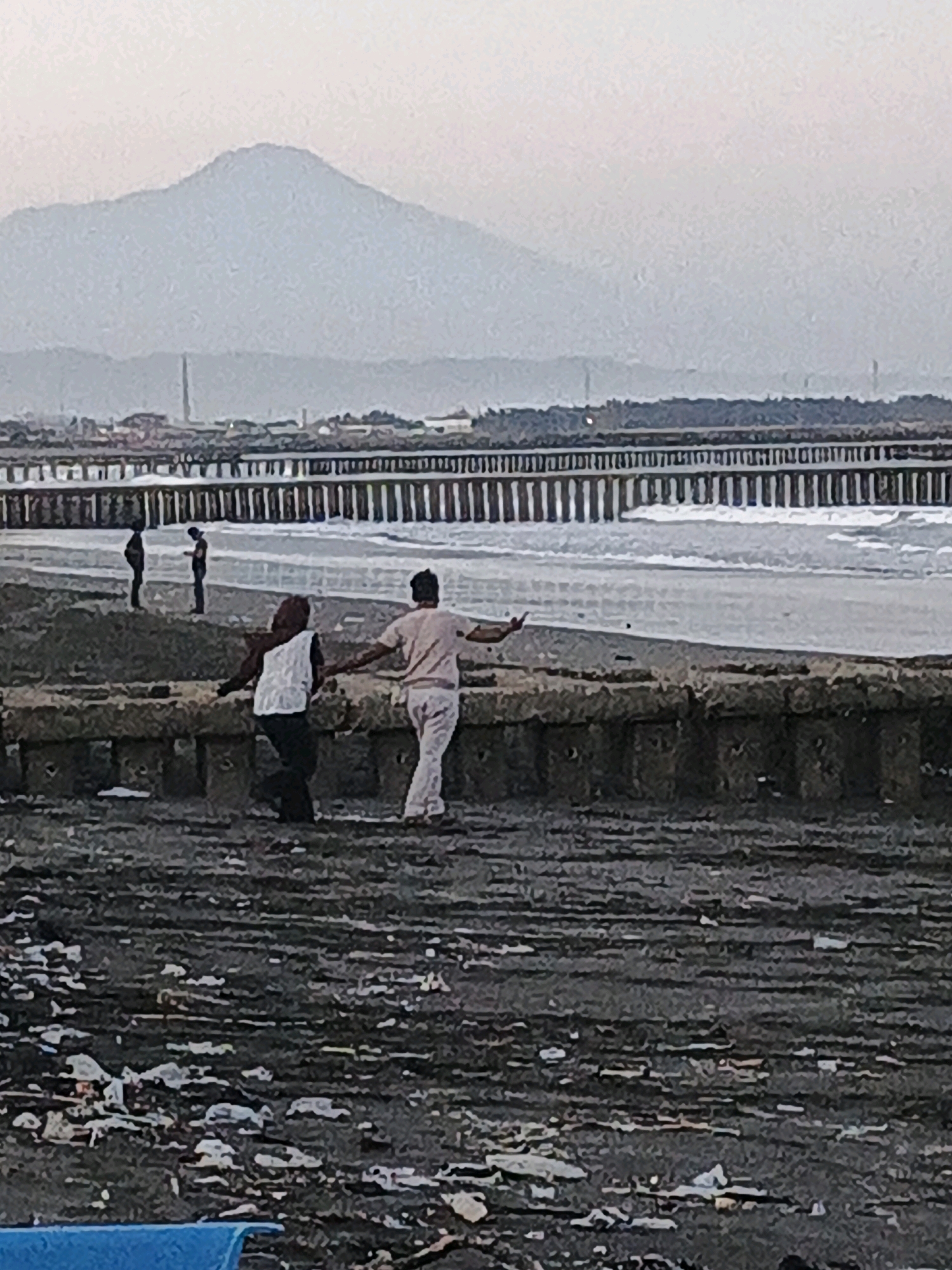 In Kelurahan Cilacap, Kecamatan Cilacap, Indonesia, a scene unfolds on a rocky shoreline under an overcast sky. In the foreground, the dark, uneven ground is littered with debris, including white plastic fragments. A stone embankment, constructed from large rectangular blocks, runs horizontally across the mid-ground. Two individuals are positioned on this embankment. One, wearing a white top and dark pants, has their back to the viewer and is looking towards the right. To their right, a second individual in a light-colored shirt and pants is facing away, with their right arm extended towards the first person. Further back, on a sandy area near the water's edge, two more figures are visible, silhouetted against the sea. One appears to be standing, while the other is slightly hunched. Beyond them, a long pier extends out into the ocean, supported by numerous vertical wooden posts. The sea is choppy, with waves breaking near the pier. In the far distance, a range of mountains forms the horizon, their peaks obscured by a hazy sky.